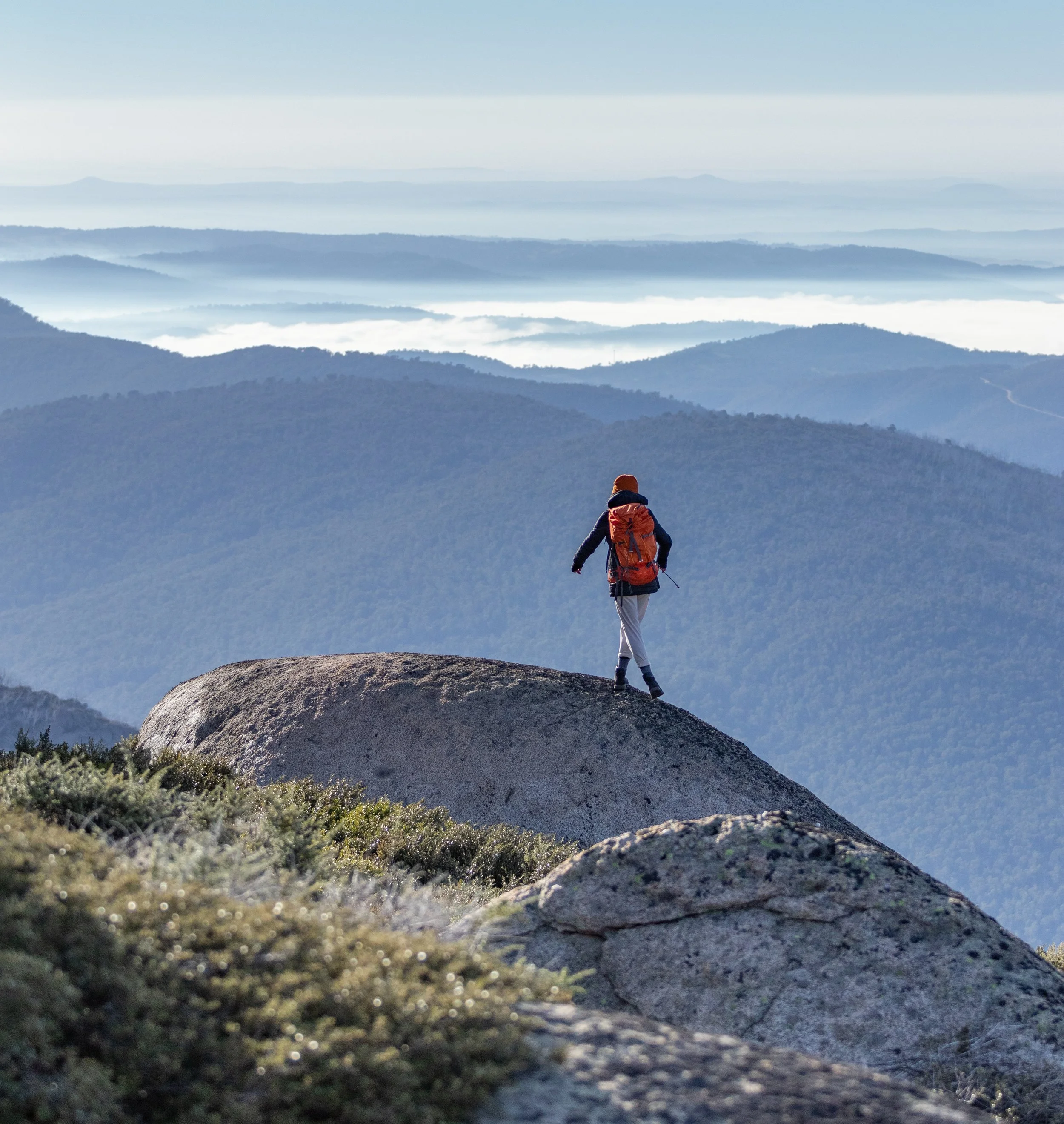 Person with a backpack hiking on a large rock on a mountain, with layers of mountain ridges and clouds in the background.