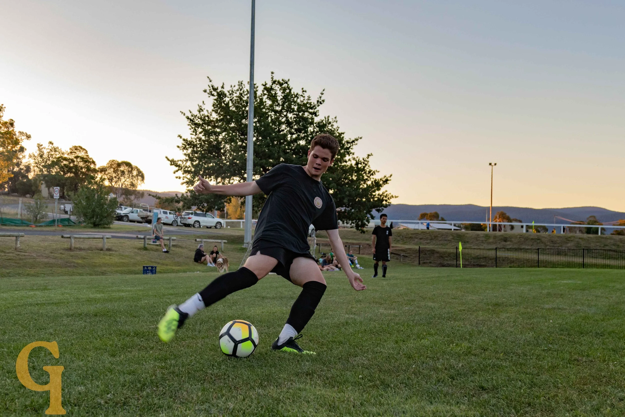 A young male soccer player in black attire and bright yellow shoes kicks a soccer ball on a grassy field during sunset, with people and cars in the background.