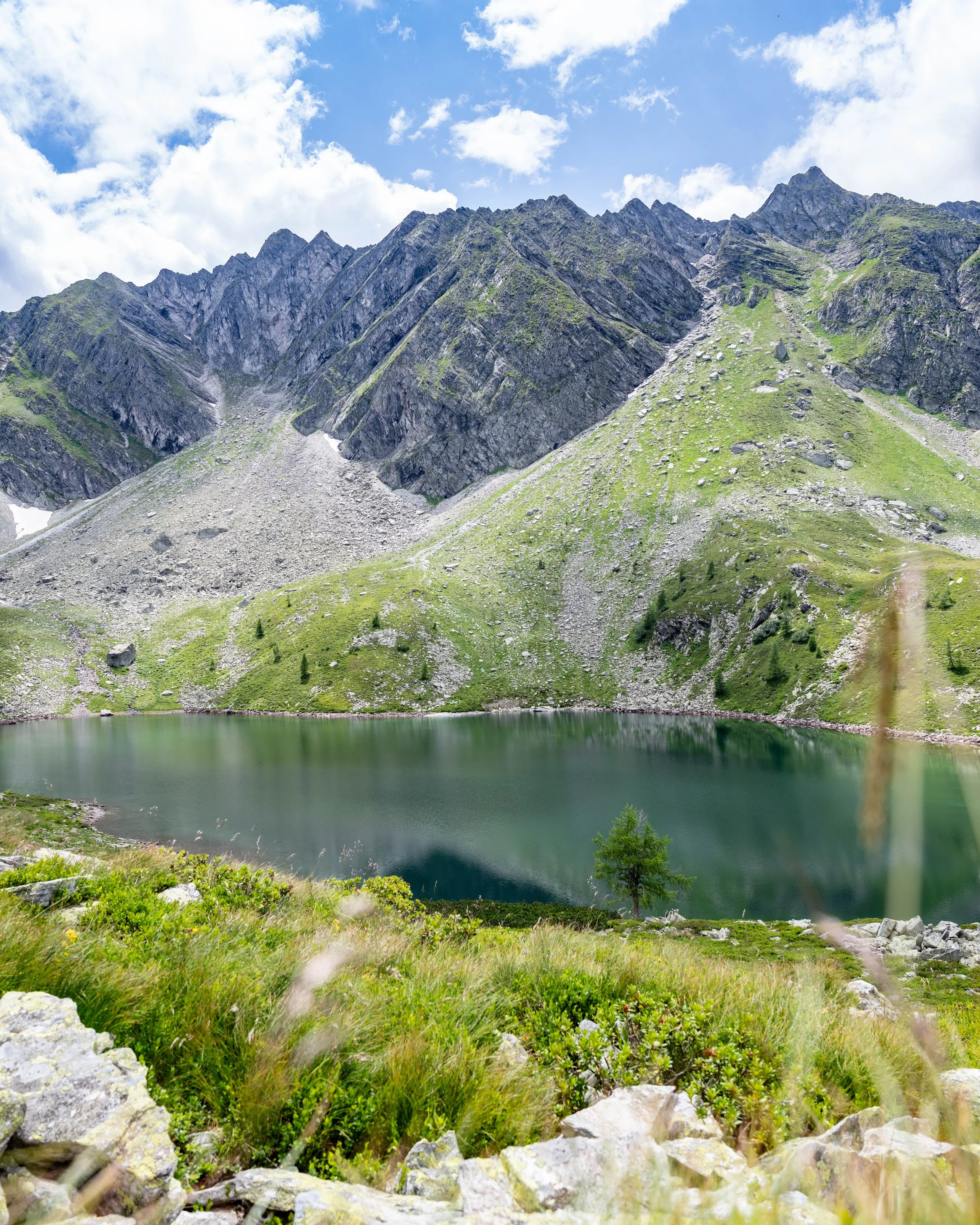 A mountain landscape with rocky peaks, green slopes, and a small lake at the base, under a partly cloudy sky.