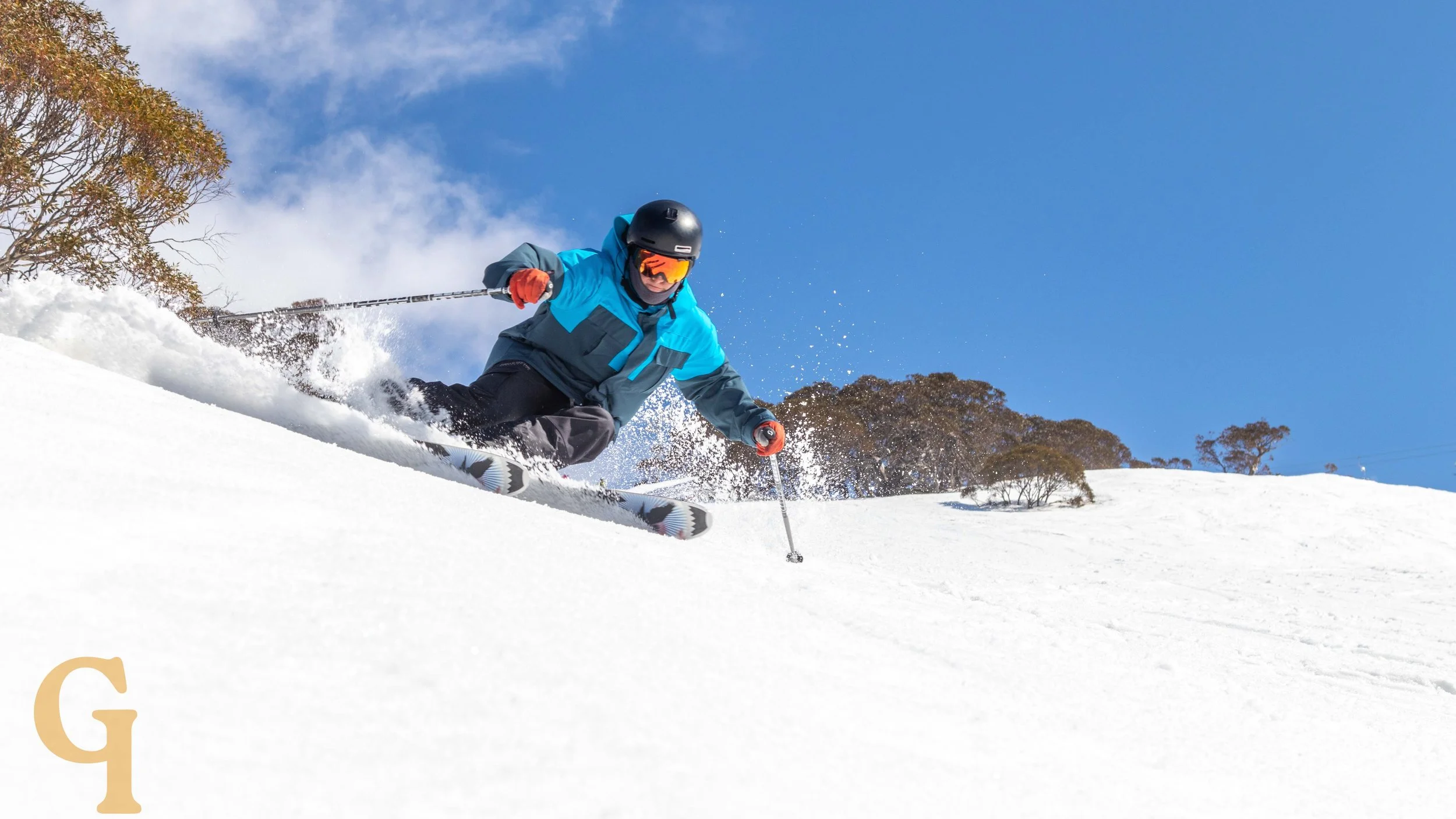 Skier in a blue jacket and black helmet skiing downhill on snow with clear blue sky and trees in the background.