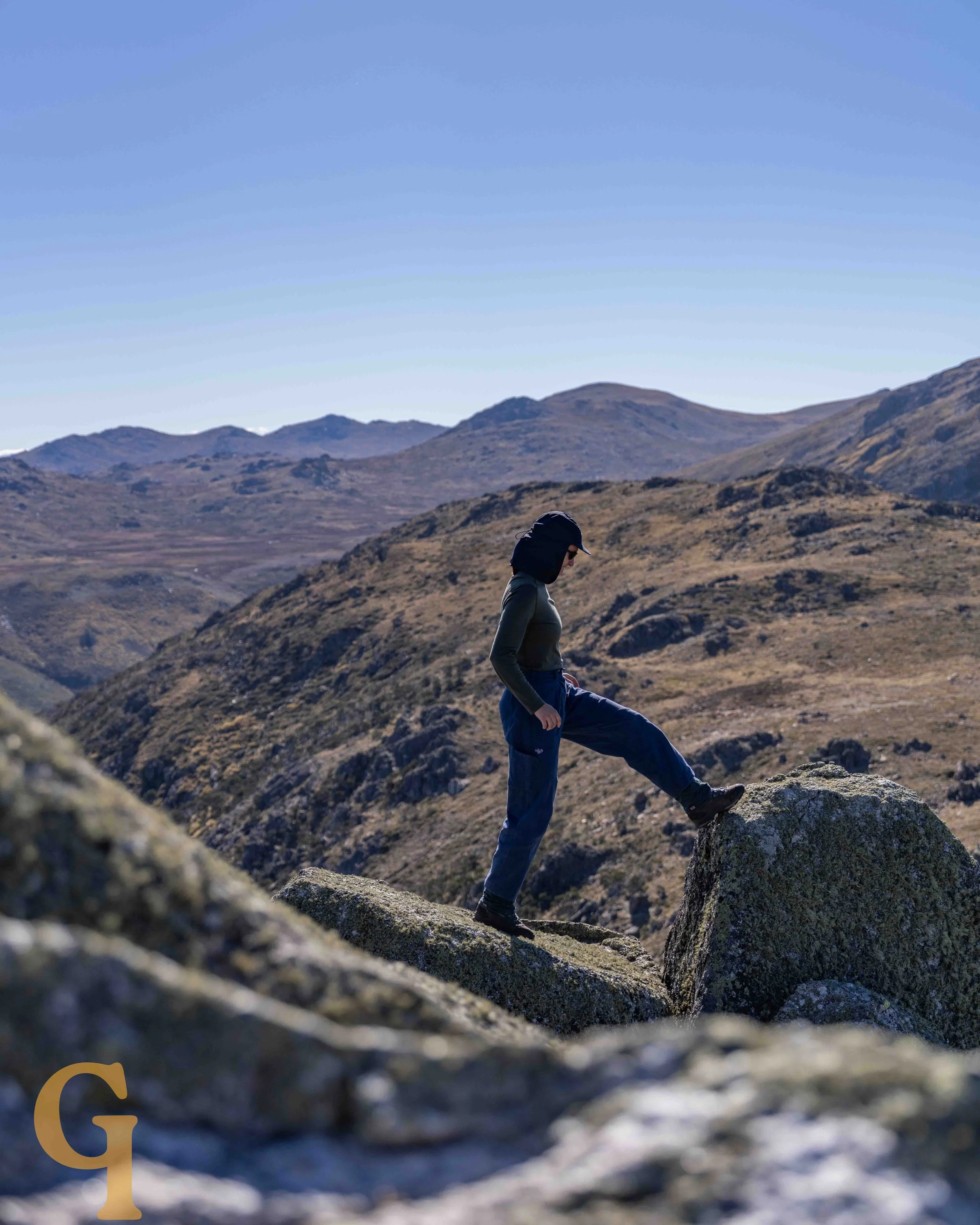 Person wearing a hat and outdoor clothing hiking on large rocks in the Snowy Mountains, NSW with rolling hills and clear blue sky.