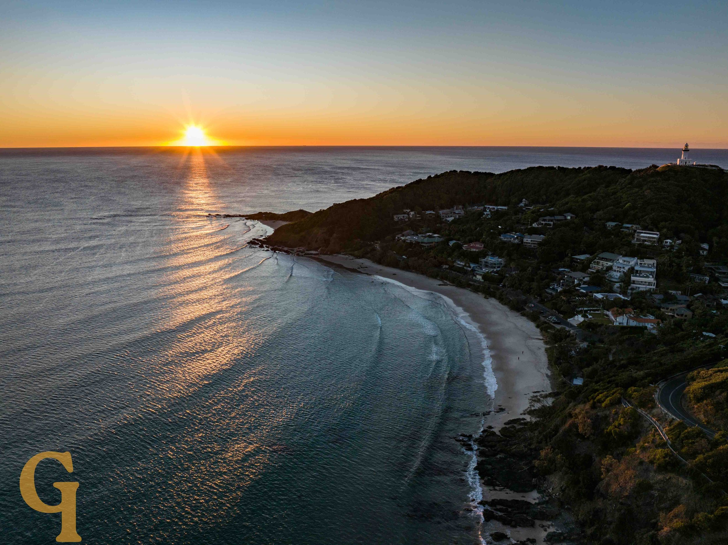 A sunrise over the ocean near a hilly coastline with trees, rocks, and a lighthouse on a distant hill.