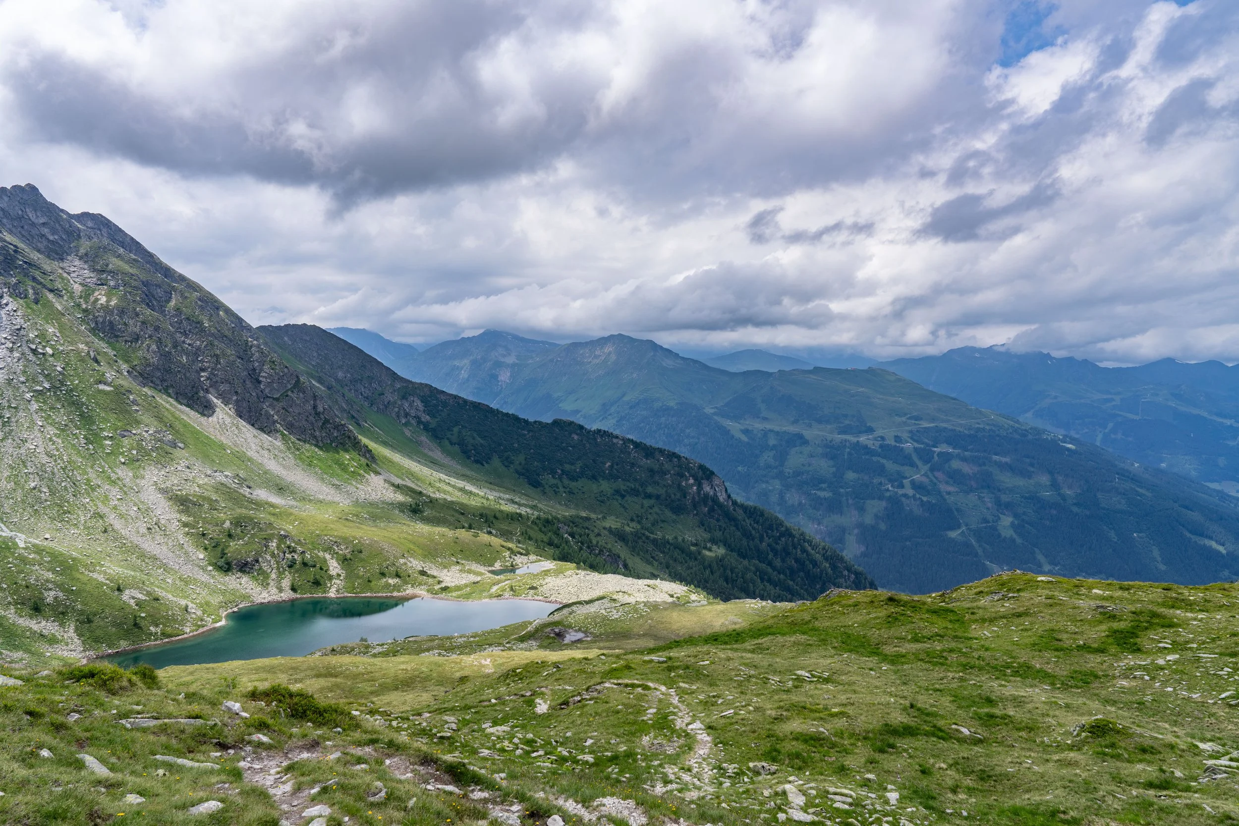 Mountain landscape with a small lake in the foreground, green rolling hills, and tall mountains under a cloudy sky.