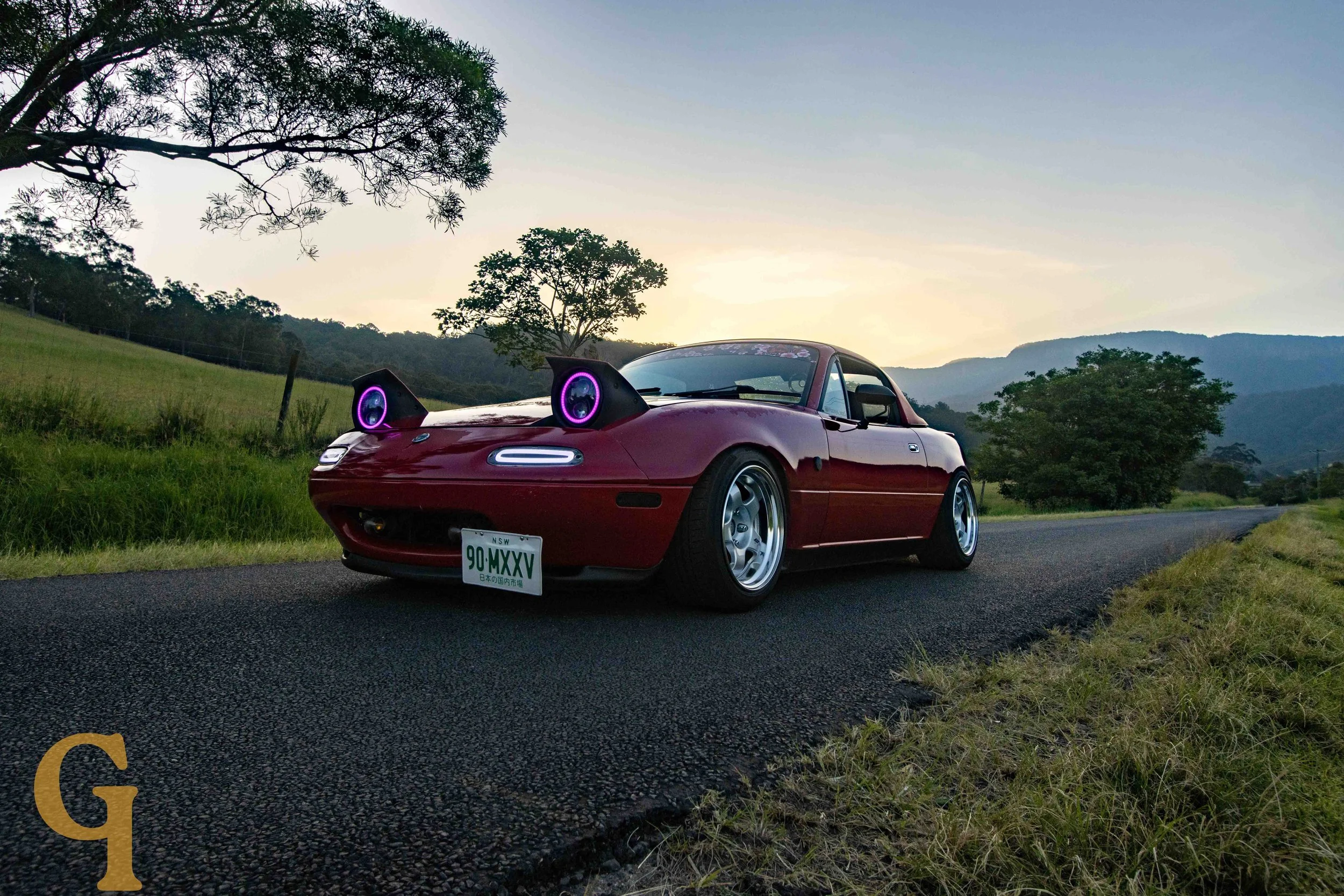 A red sports car with modified headlights emitting purple light parked on a rural road with green grass, trees, and mountains in the background during sunset.
