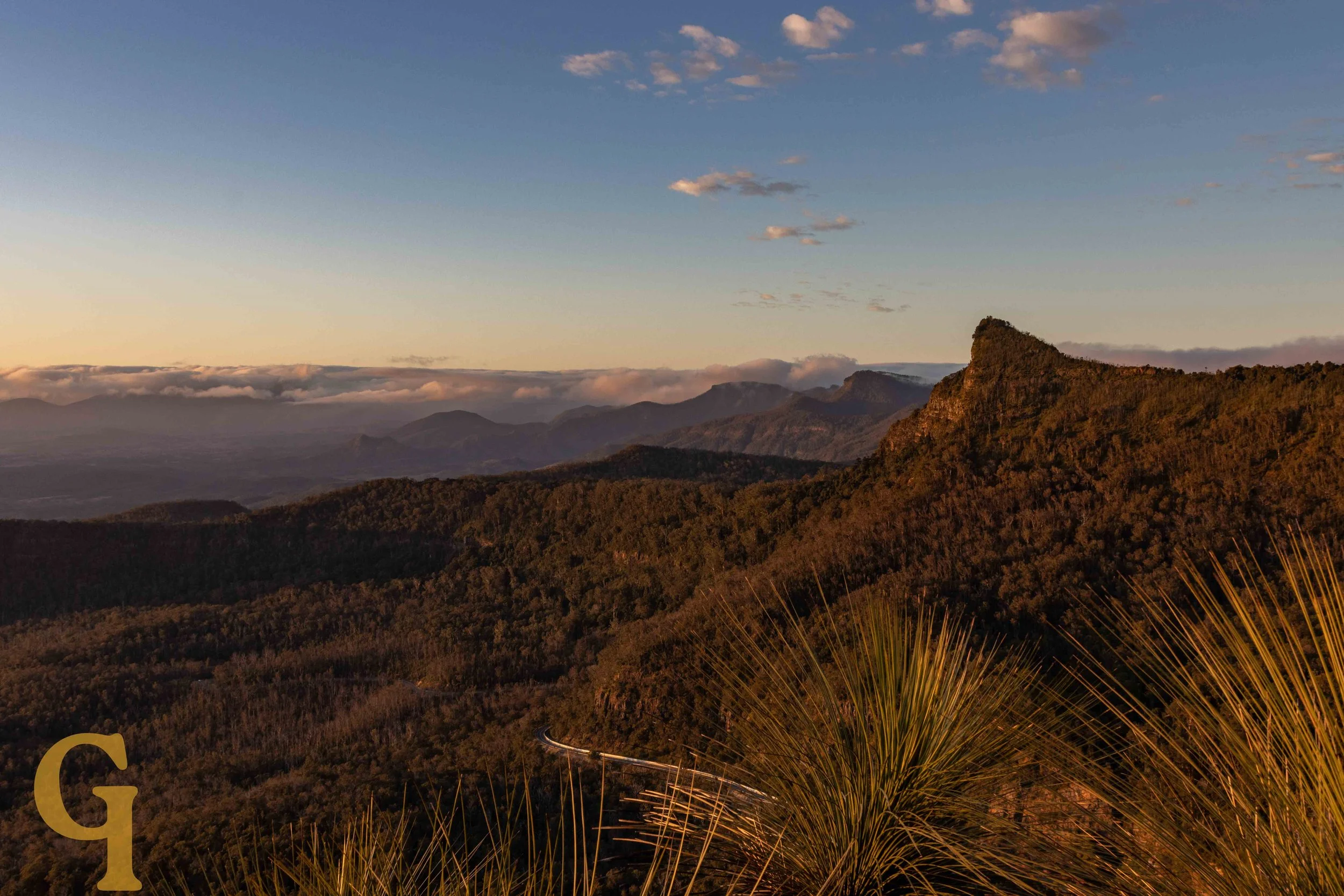 Sunrise over a mountain range with a winding road visible near the bottom and a clear sky with some clouds.