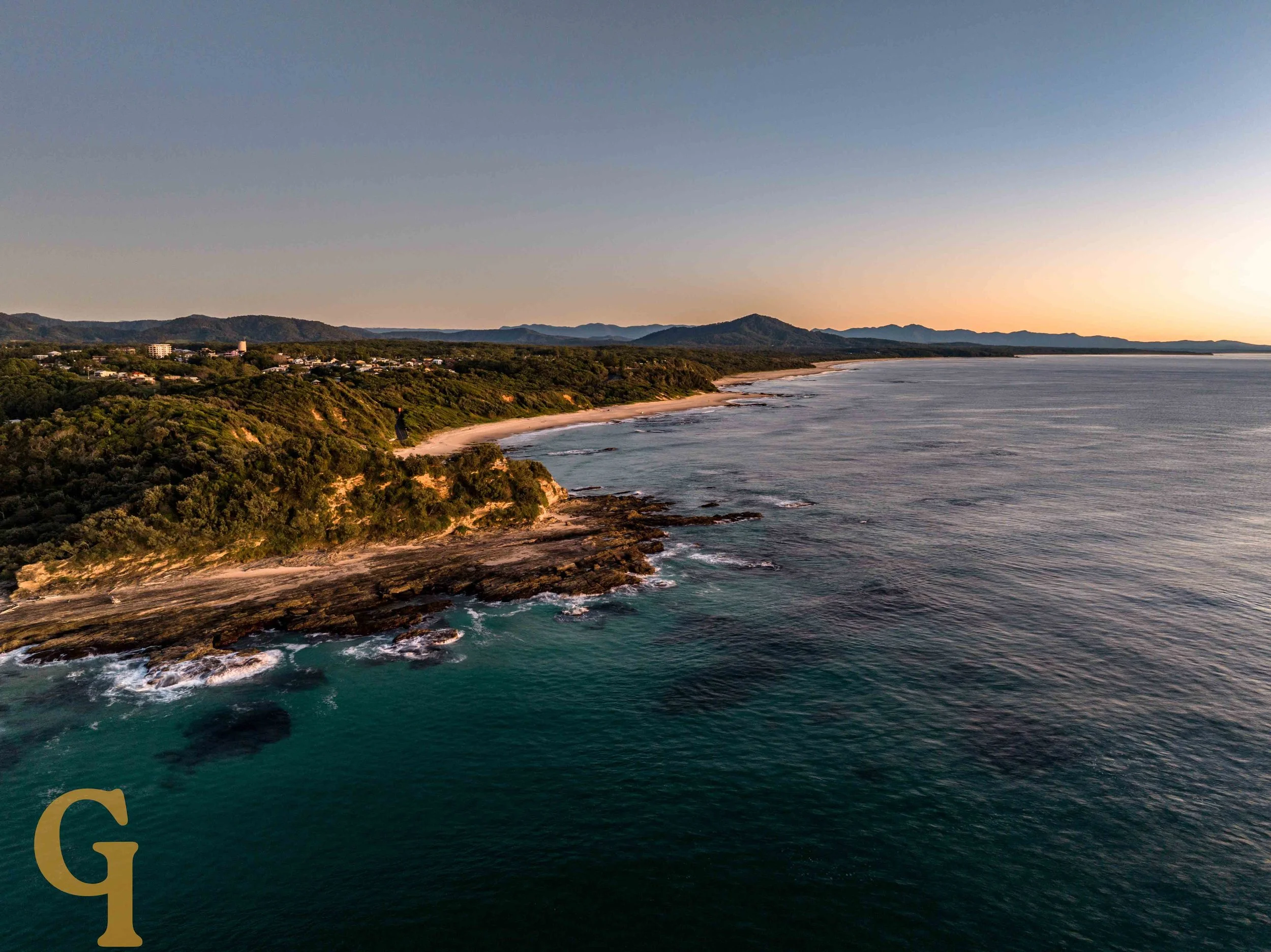 Aerial view of a coastline with rocky shores, sandy beaches, green forests, hills, and distant mountains at sunrise.