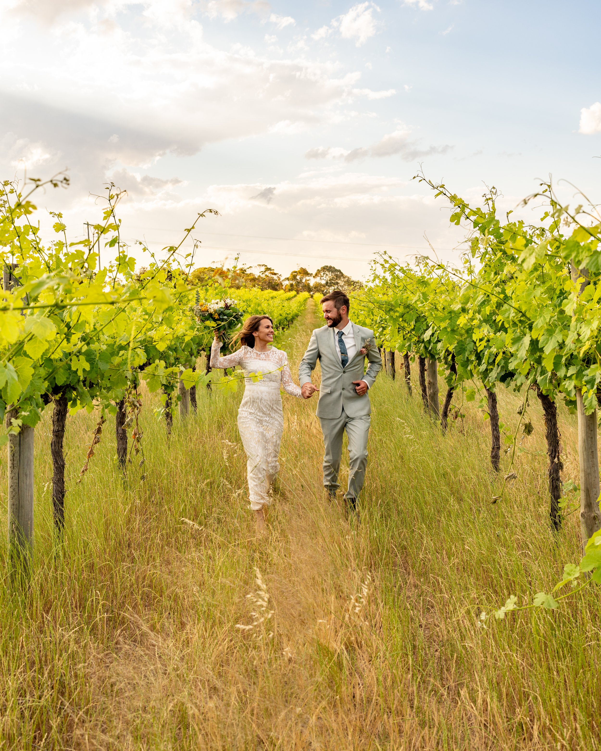 A bride and groom walking hand-in-hand through a vineyard on their wedding day, with the bride holding a bouquet and smiling at each other on a sunny day.