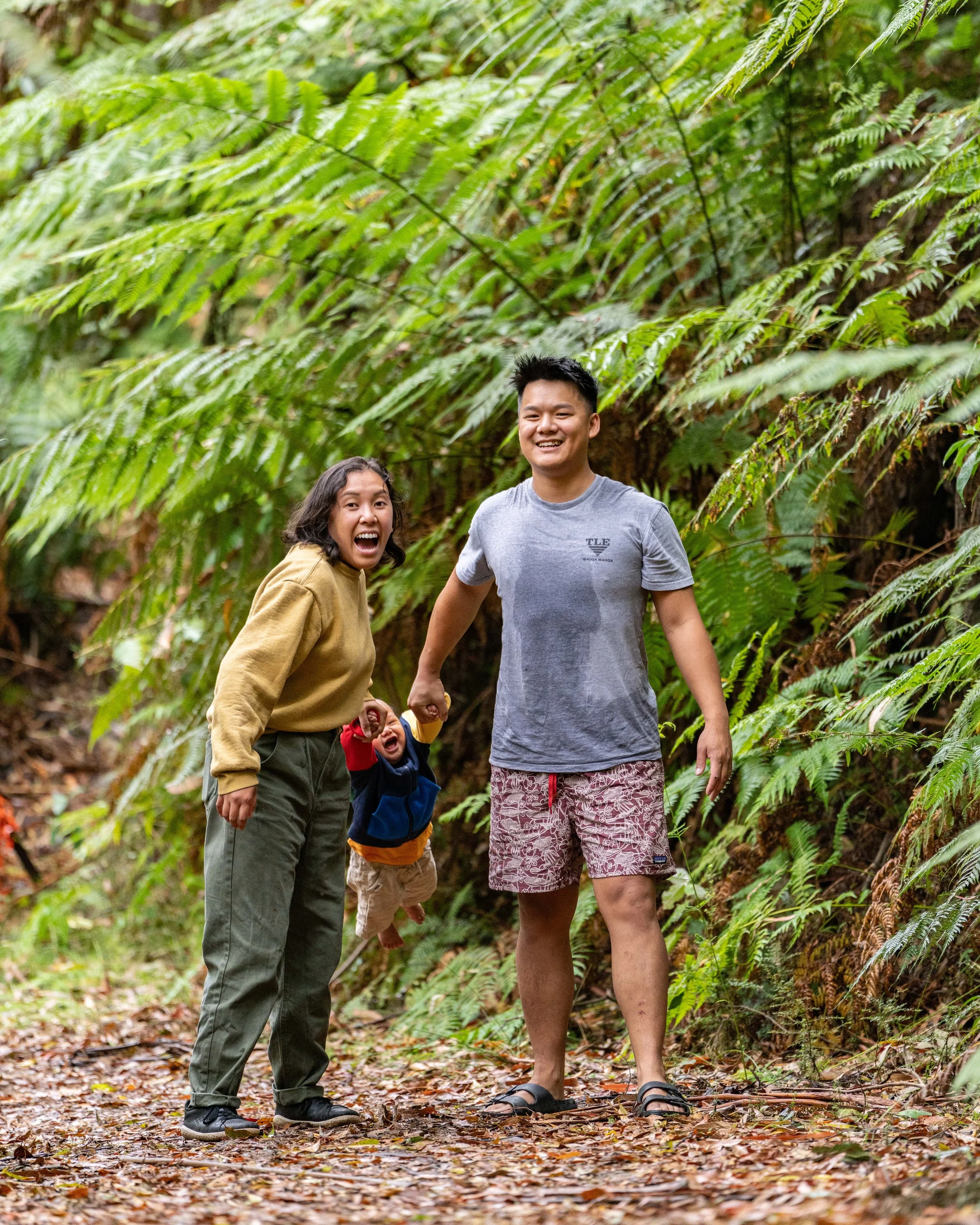 A family of three, including a woman, a man, and a toddler, enjoying a walk through a lush green rainforest. The woman and man are smiling and the toddler is being held by the woman. Ian Grant is skilled in a variety of portrait photography.