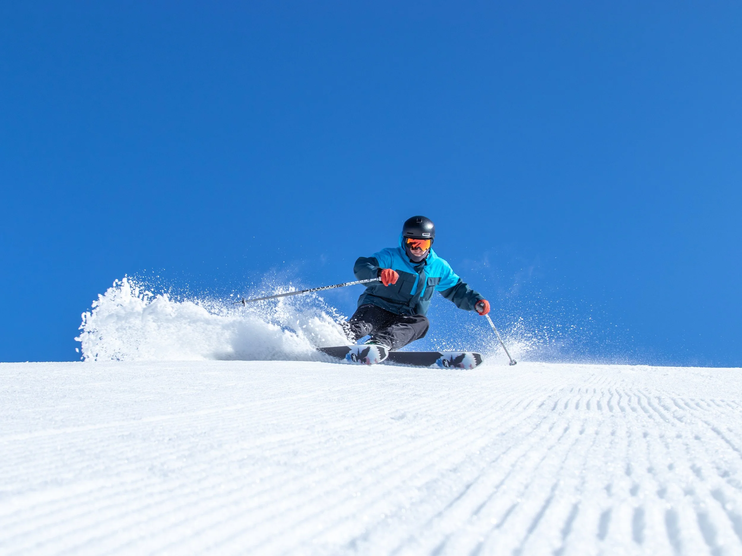 A skier wearing a blue jacket, black helmet, and goggles skiing on snow with a clear blue sky in the background.