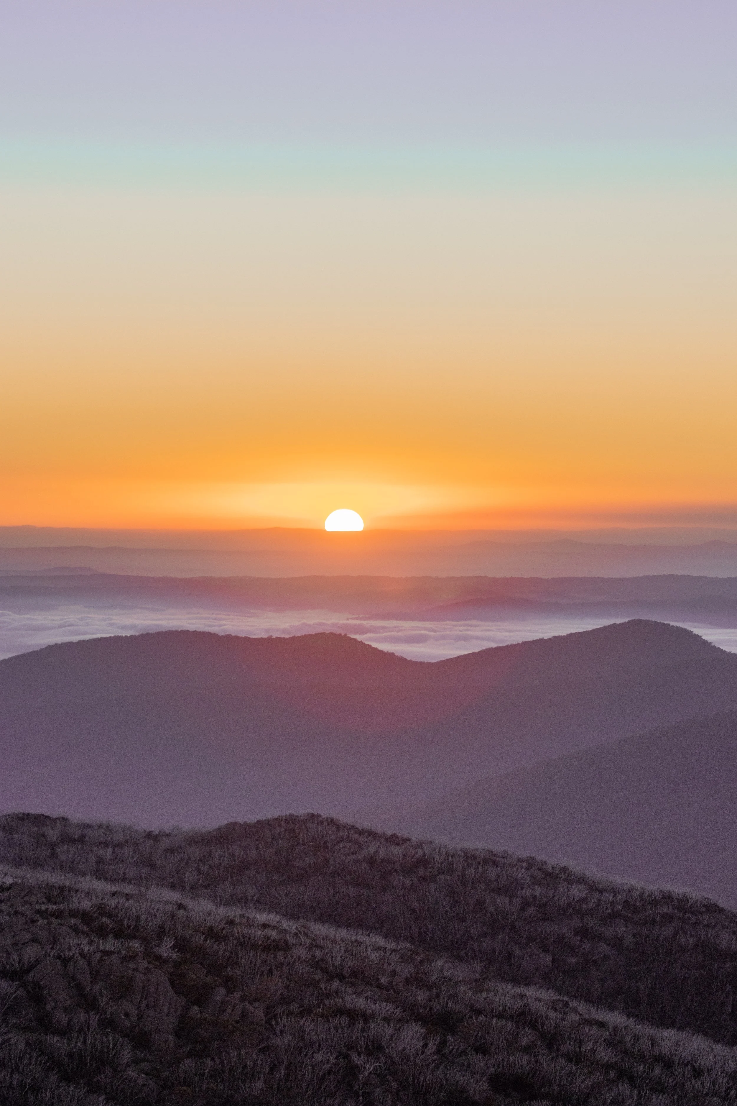 Sunset over mountain range with a colorful sky filled with orange, pink, and purple hues, and layers of mist or clouds in the valleys between the hills.