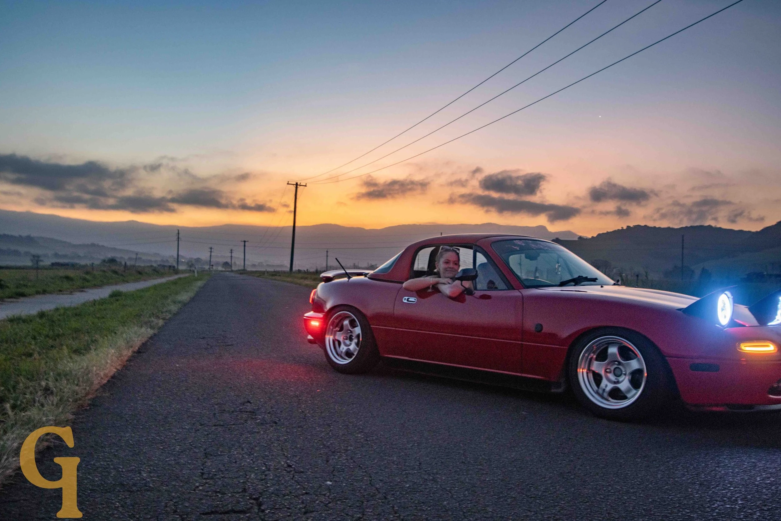 A woman smiling and leaning out of the driver's side window of a red sports car on a rural road at sunset with hills and power lines in the background.