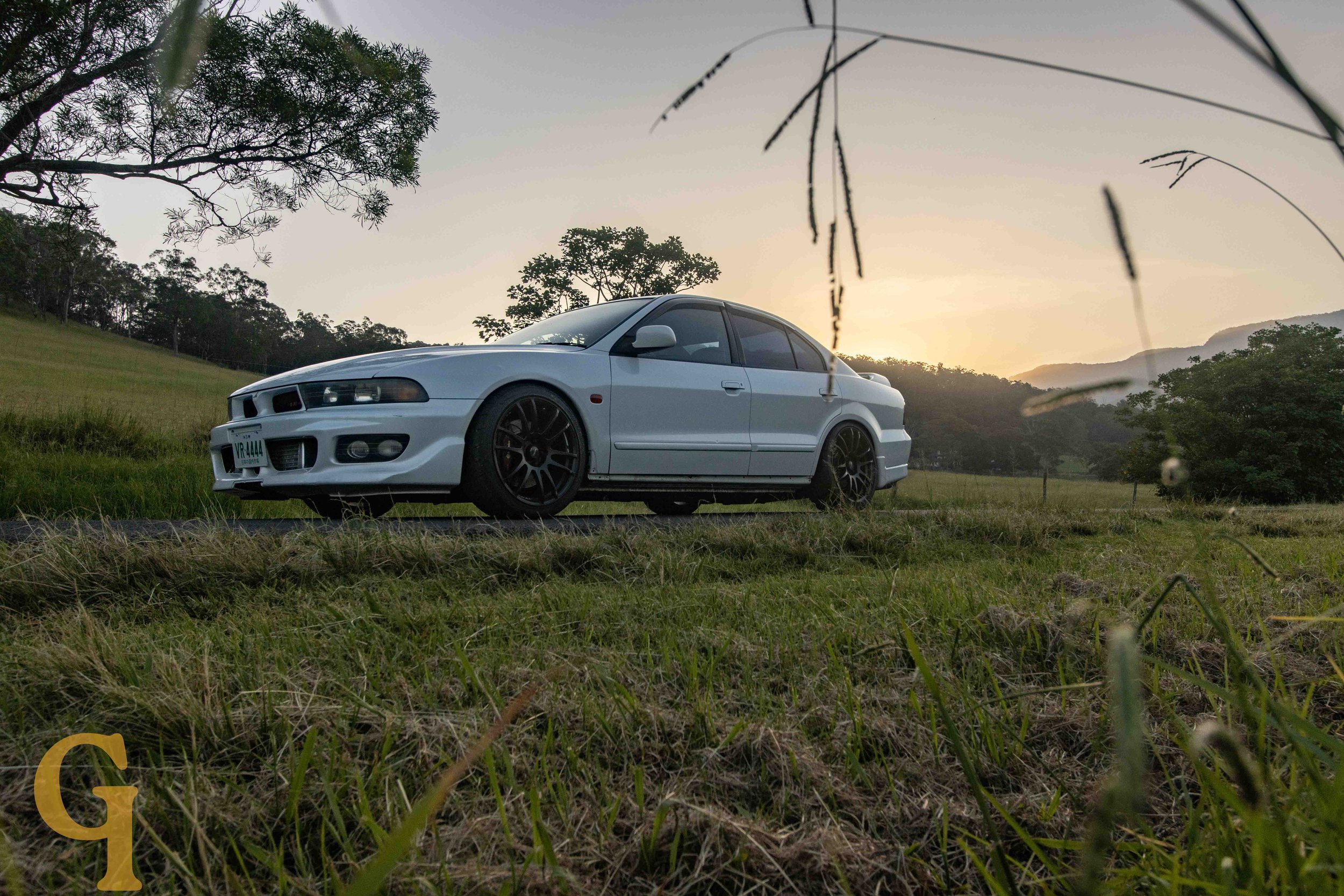 A white sedan car parked on grass with trees and hills in the background during sunset.