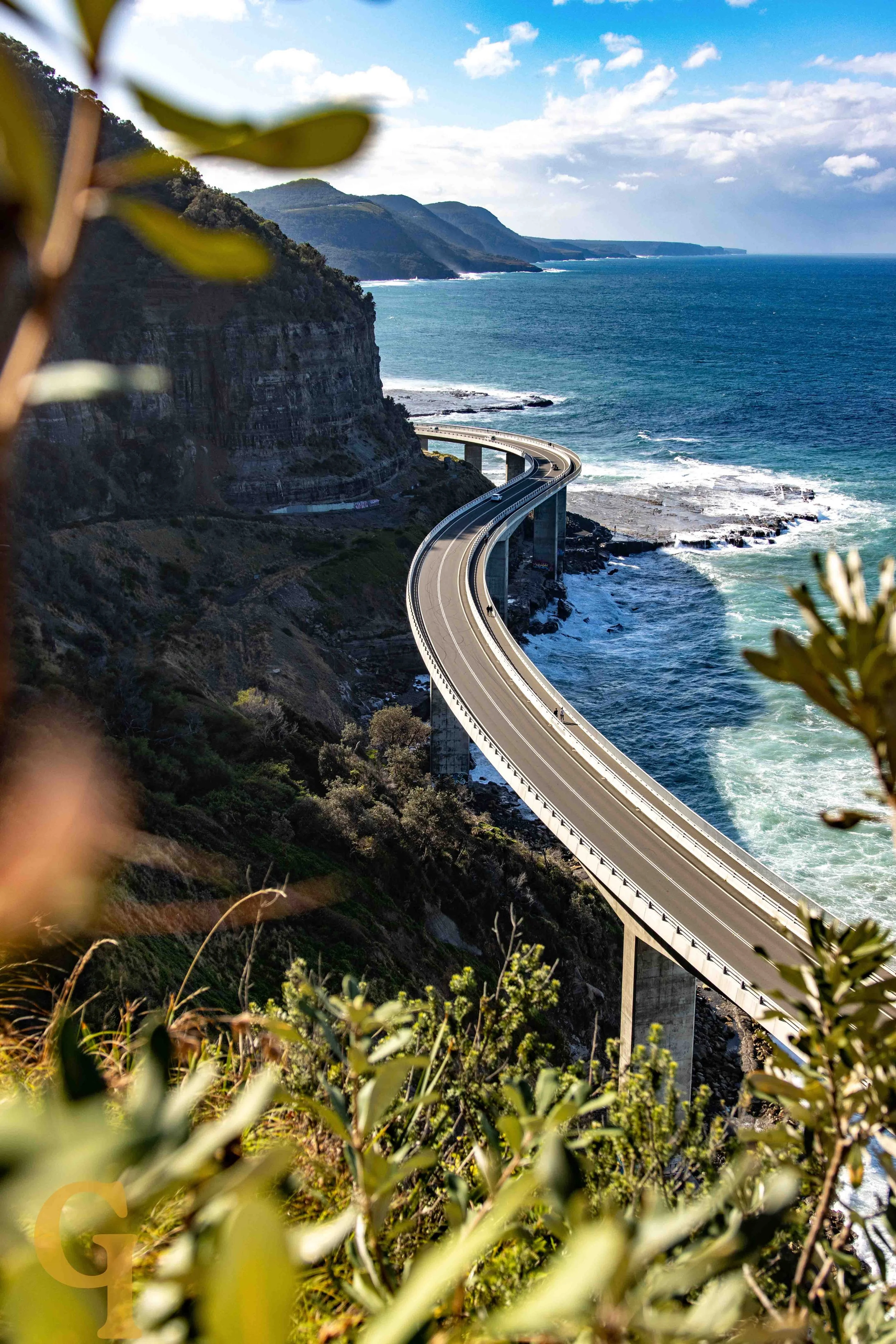 A coastal highway bridge winding along cliffs with the ocean below and green mountains in the background.