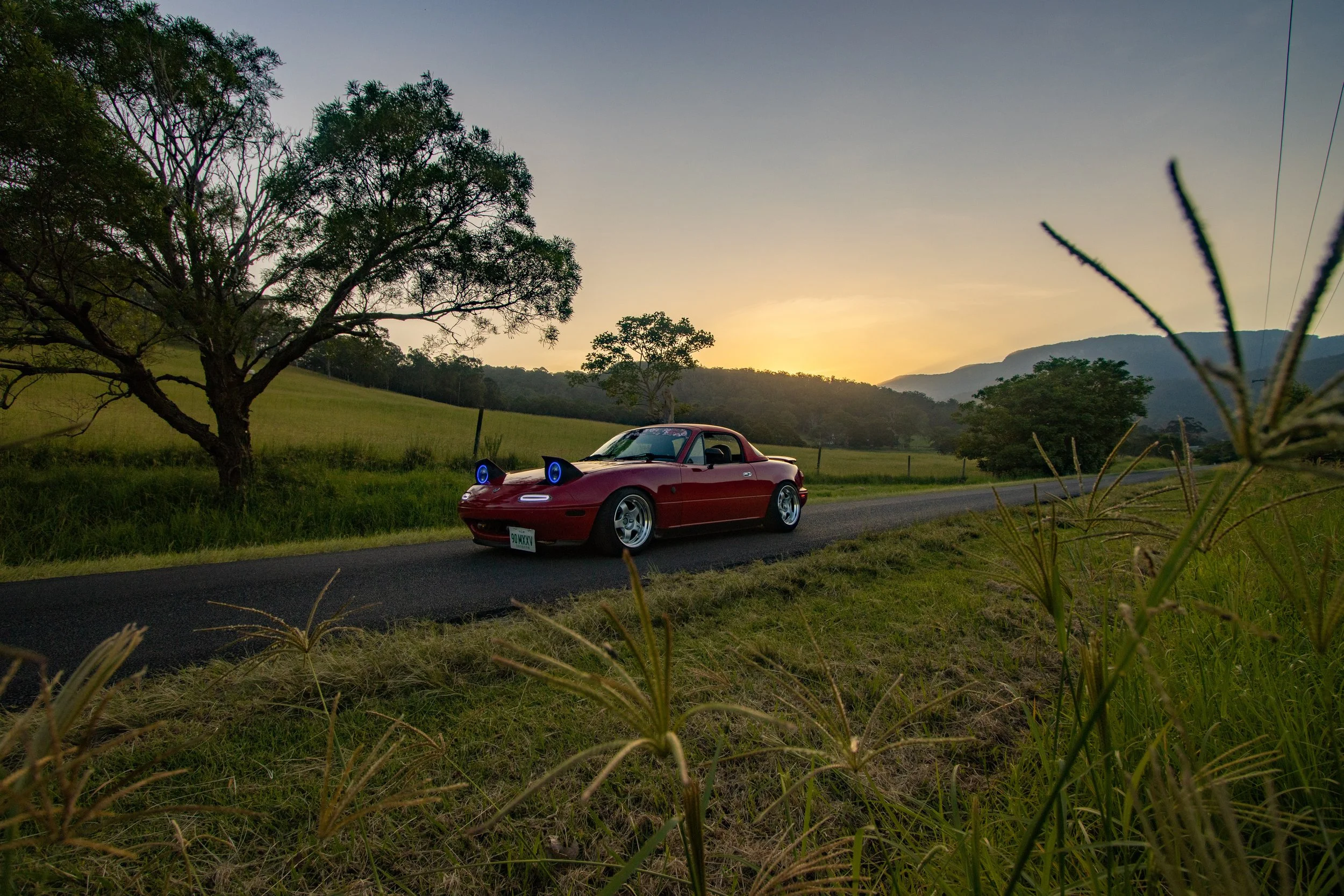 A red Mazda MX-5 Miata with cat ears and glowing blue eyes headlights, parked on a country road at sunset, with trees and hills in the background.