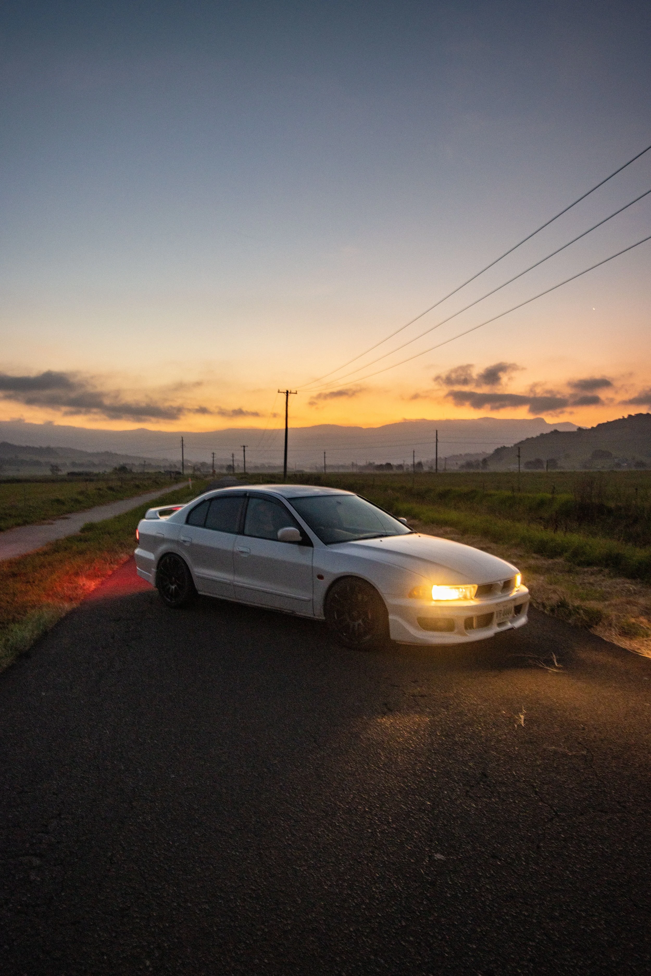 White sedan car with illuminated headlights parked on side of gravel road at sunset in rural landscape.