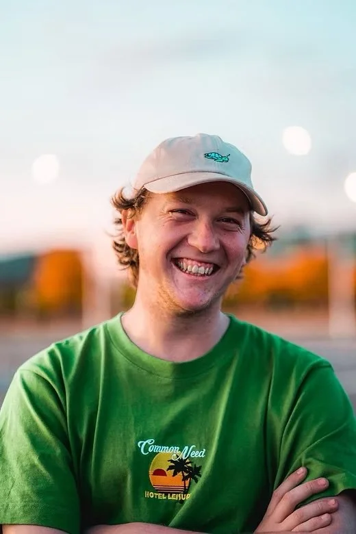 A young man smiling outdoors at sunset, wearing a beige cap with a small logo and a bright green T-shirt with a logo reading 'Common Need Hotel Leisur' and a sunset graphic with palm trees.