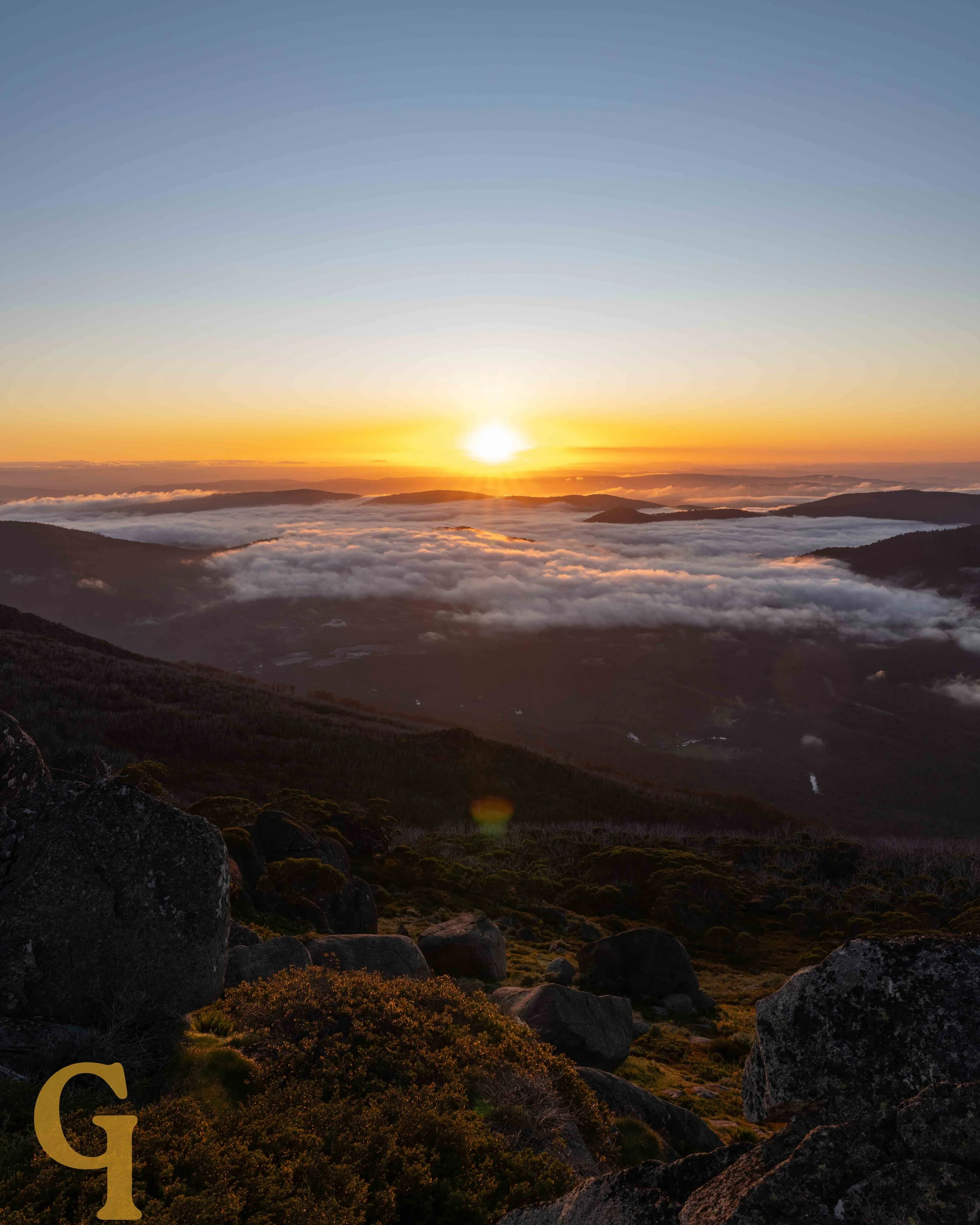 Sunrise over mountain landscape with clouds, rocky foreground, and rolling hills in distance.