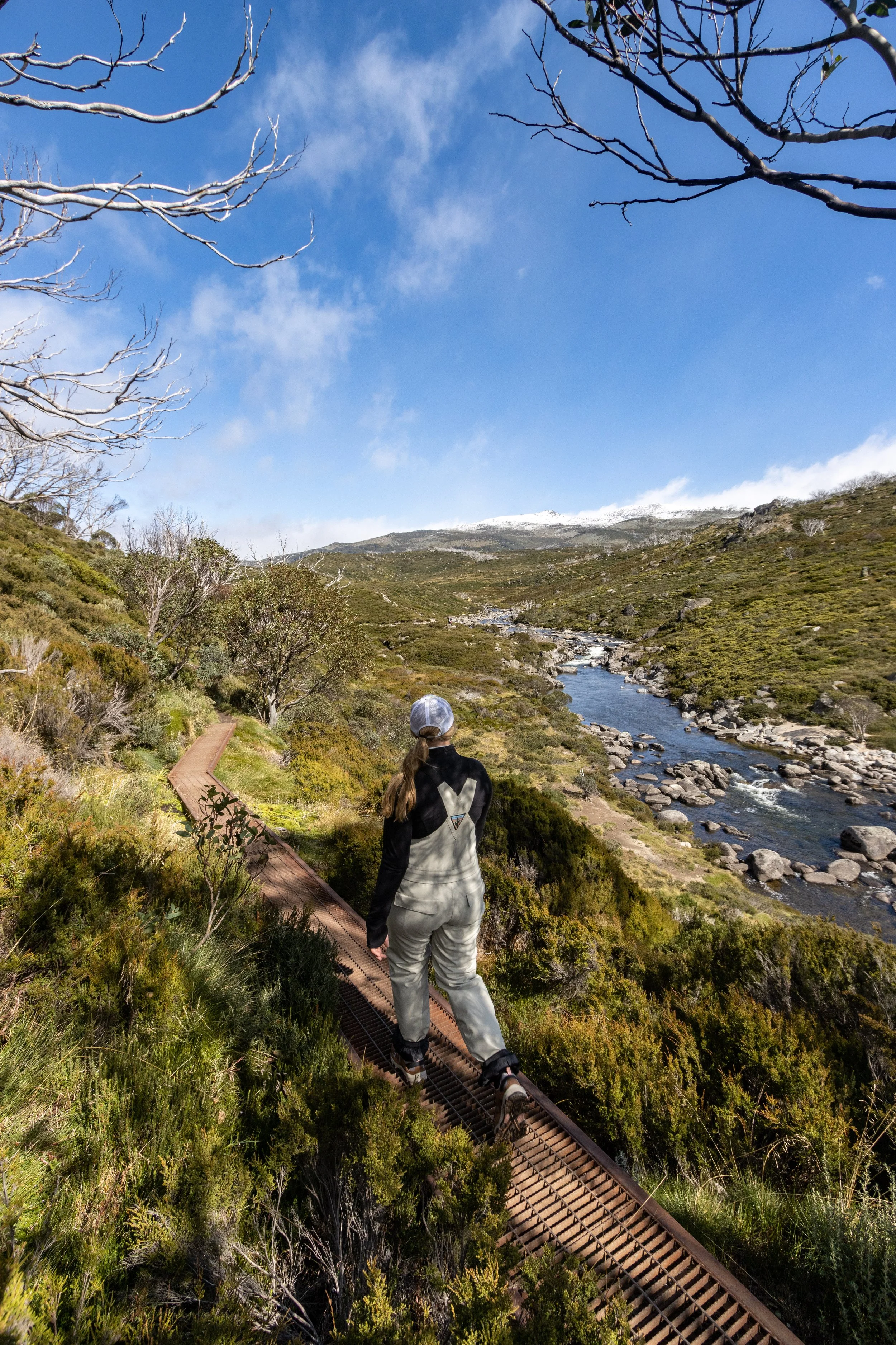 A person walking on a narrow, rusty metal railway track through green shrubland near a flowing river with rocks, under a partly cloudy blue sky.