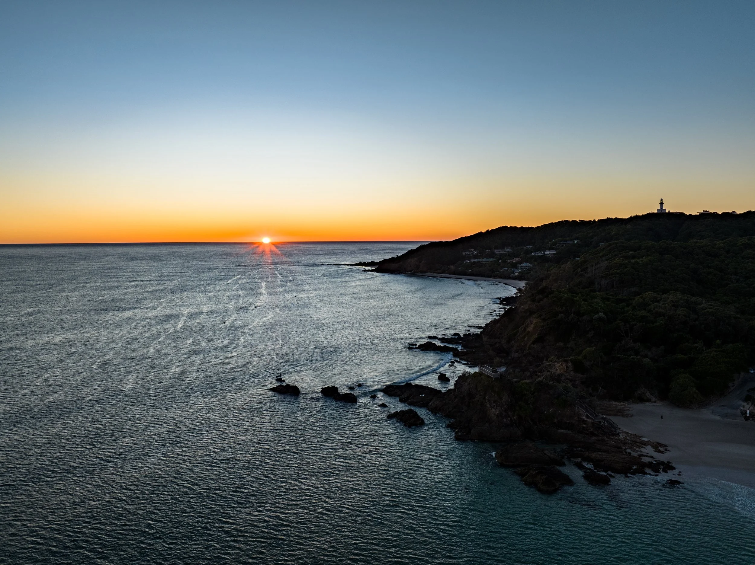 A sunset over the ocean near a hilly coastline with trees, rocks, and a lighthouse on a distant hill.