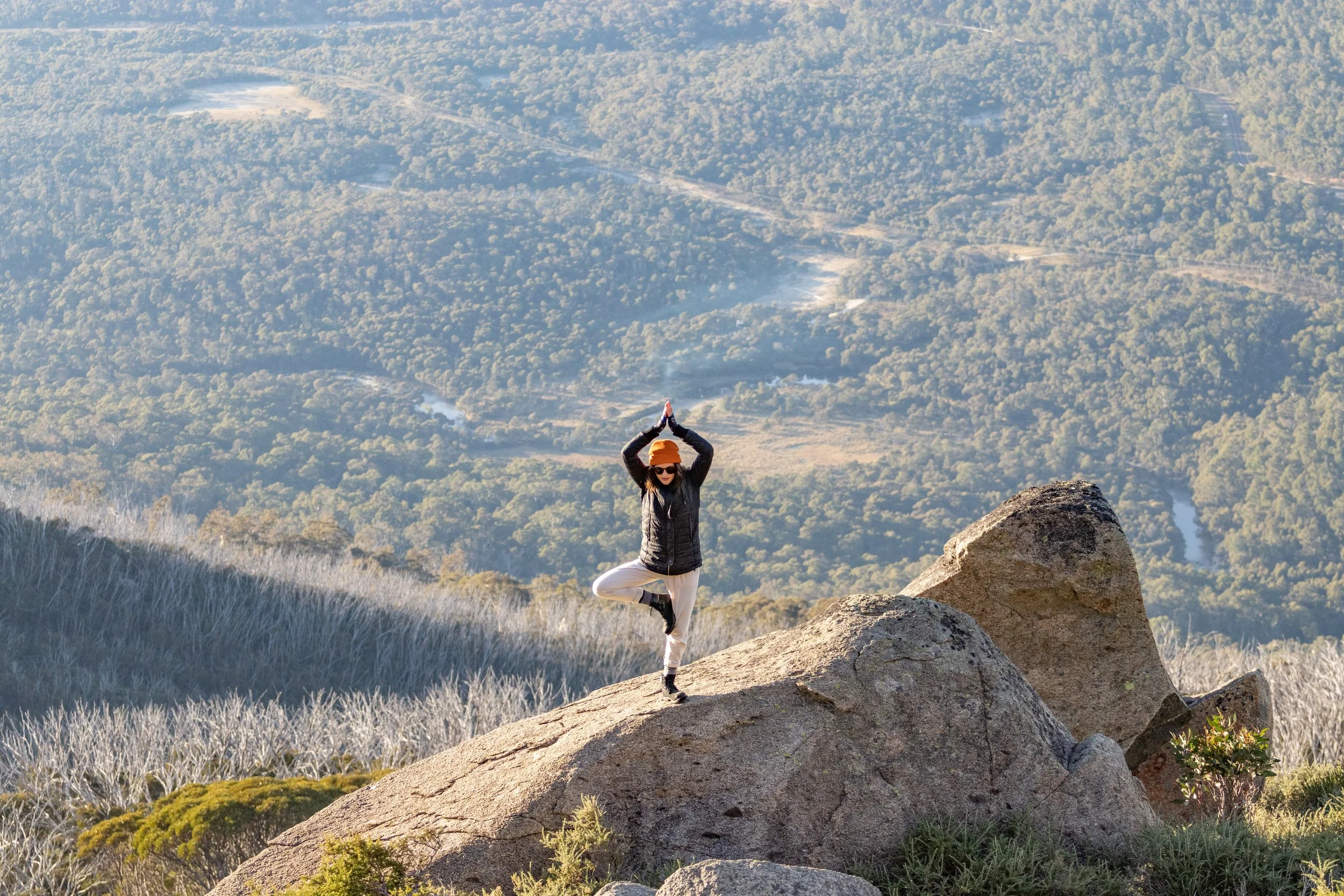 A person practicing yoga on a large rock in a mountainous landscape during daytime, with a river visible in the background.
