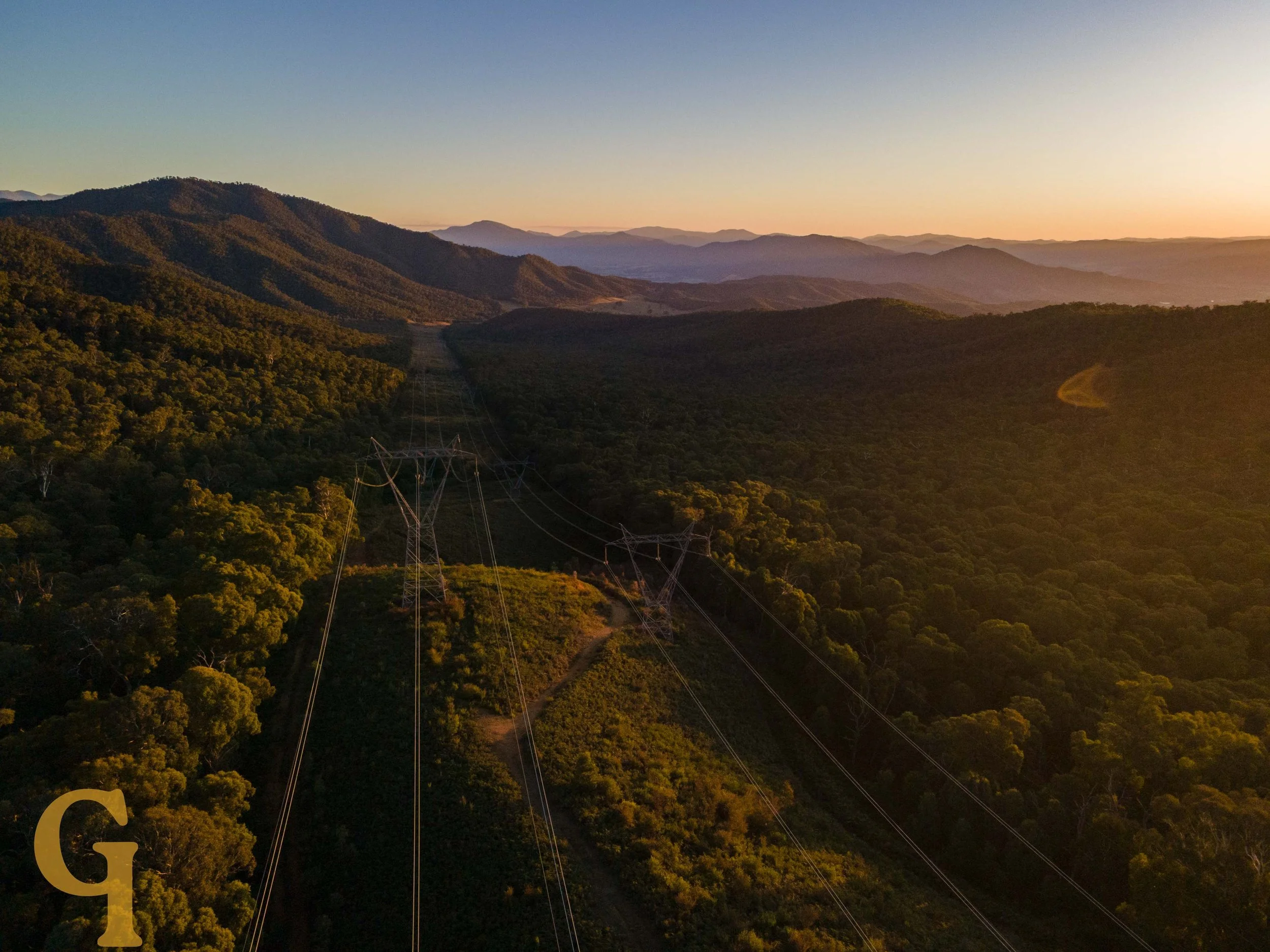 Aerial view of a forested mountainous landscape at sunset with power lines running through the hills.