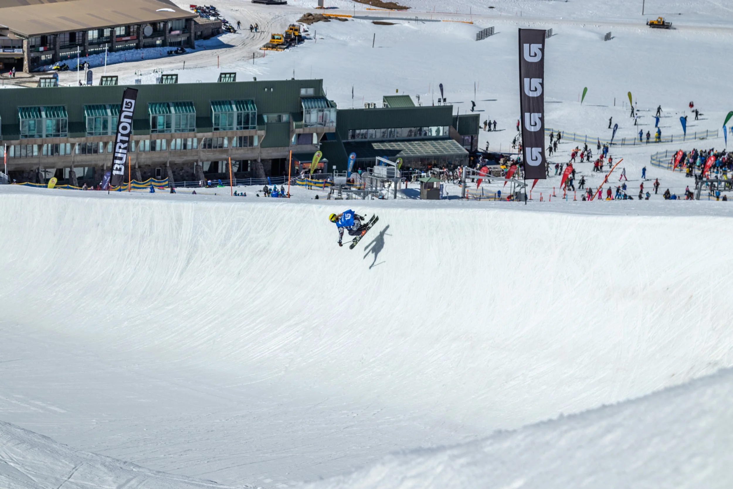 A snowboarder mid-air performing a trick on a snowy halfpipe at a ski resort.