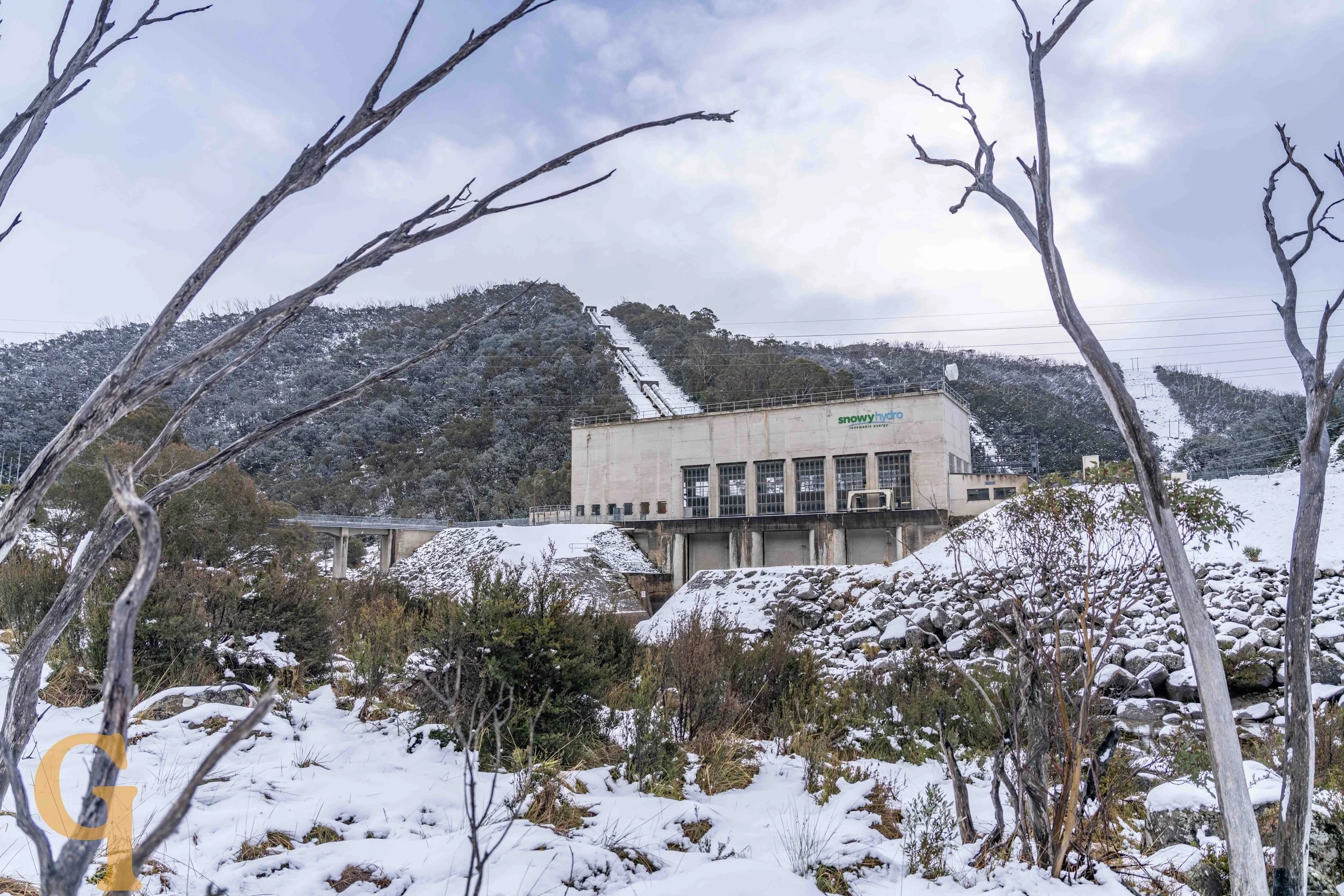 Snow-covered landscape with a hydroelectric dam at the base of a mountain, leafless trees in the foreground, and ski slopes visible on the mountain above.