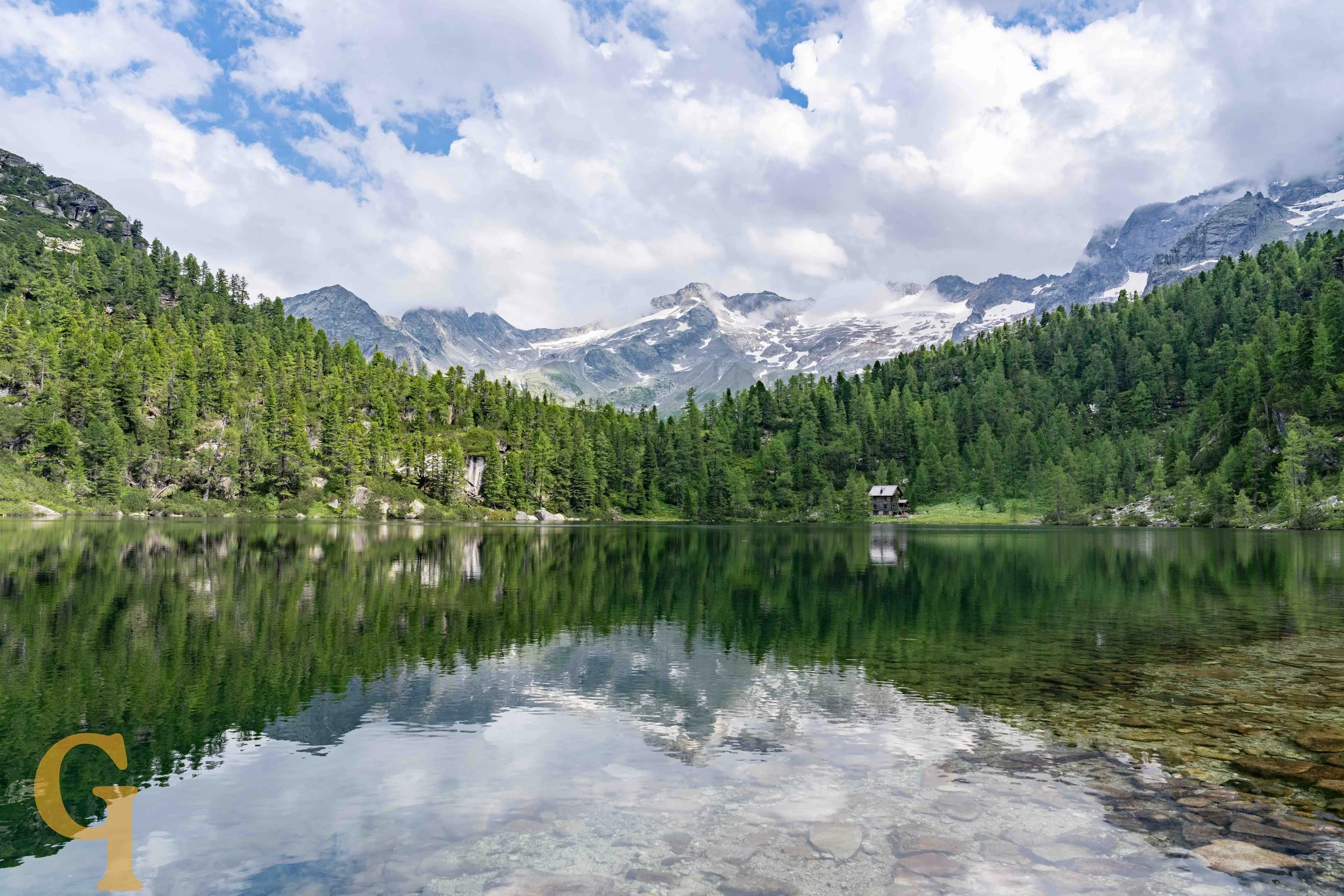 Scenic view of a mountain lake surrounded by dense green trees with snow-capped mountains in the background under partly cloudy sky.