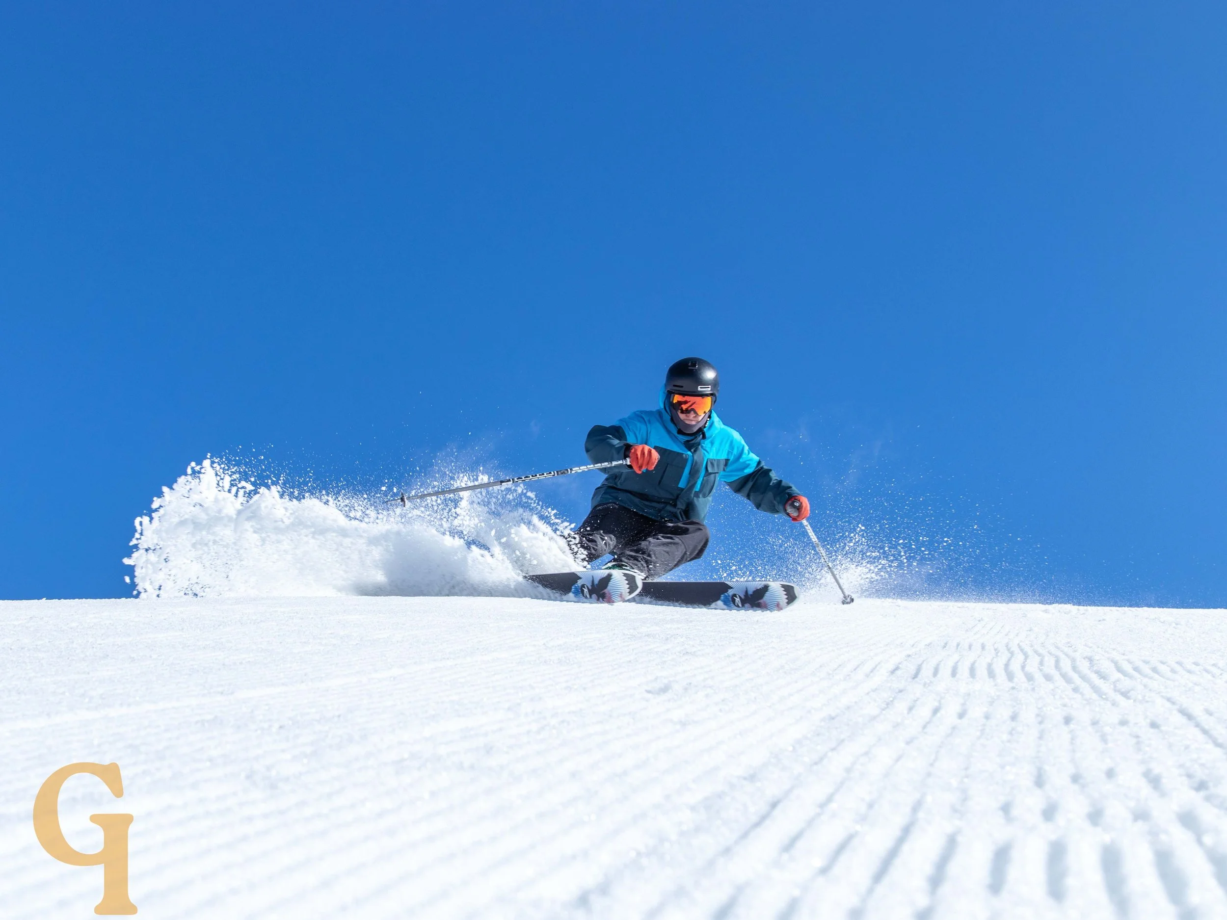 A skier wearing a blue jacket, black helmet, and goggles skiing on snow with a clear blue sky in the background.