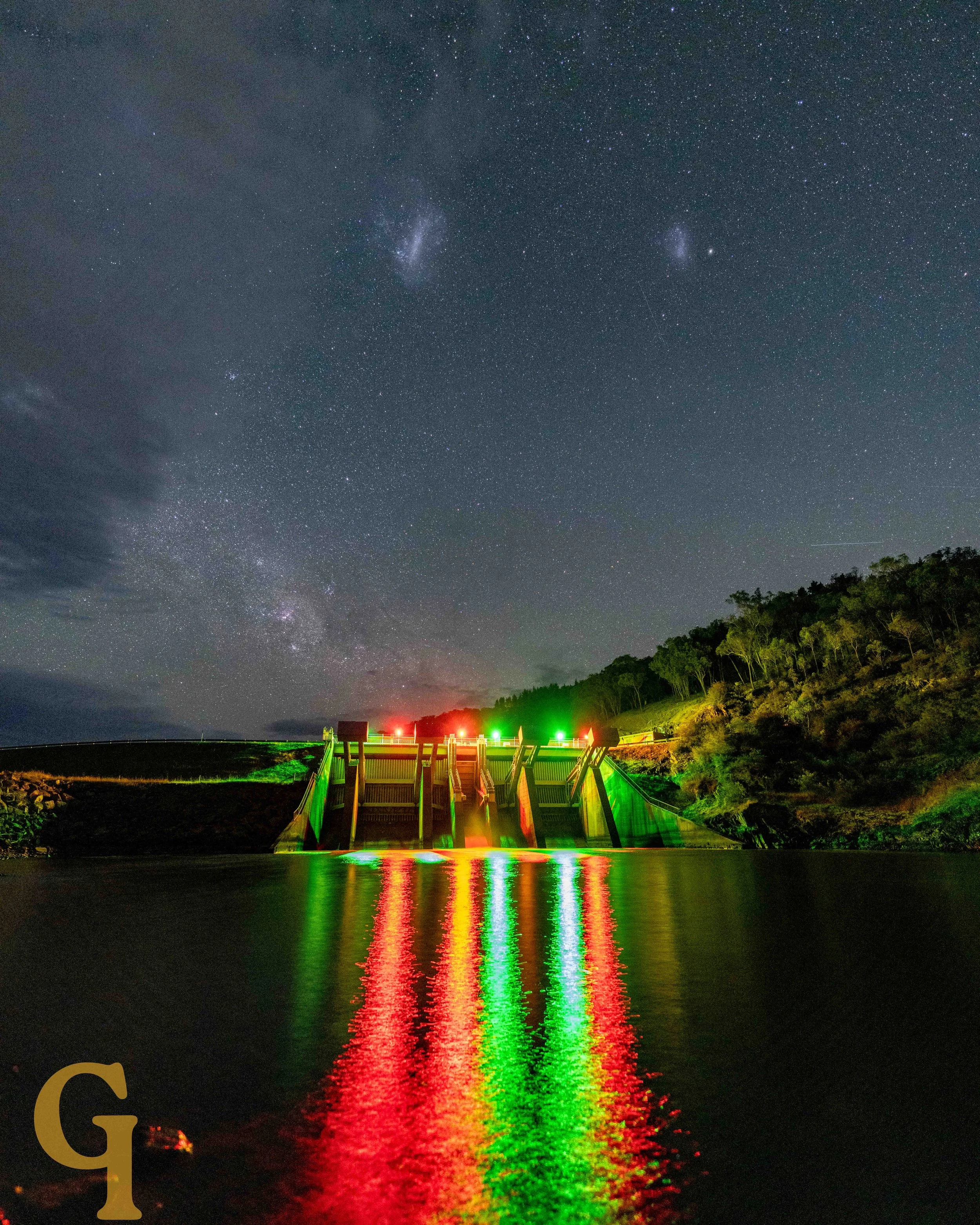 Nighttime scene of a dam with water releasing, illuminated with red, green, and white lights, reflecting on the water, under a starry sky with the Milky Way galaxy visible.
