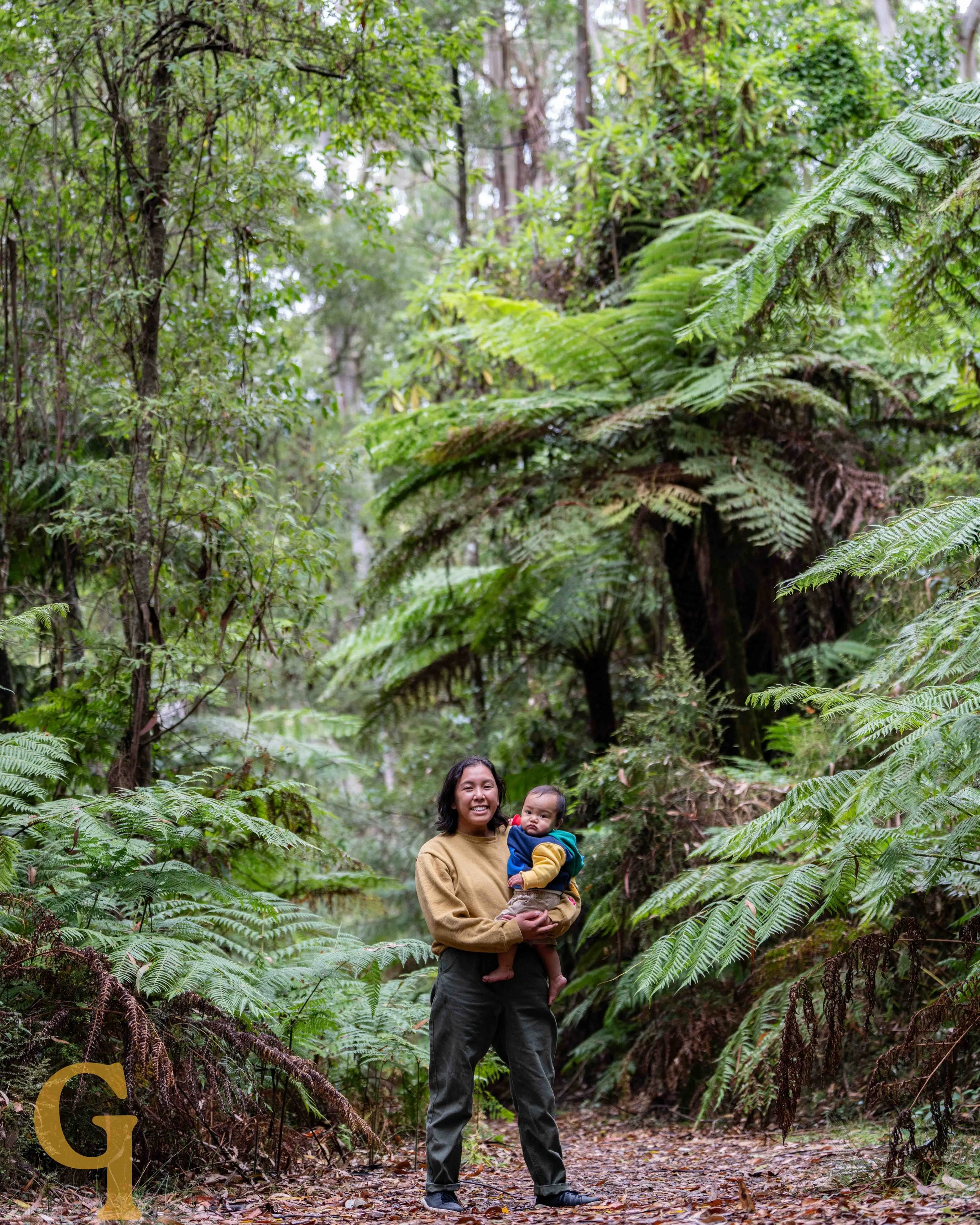 A woman is standing in a lush, green forest holding a baby. Both are smiling and appear happy.