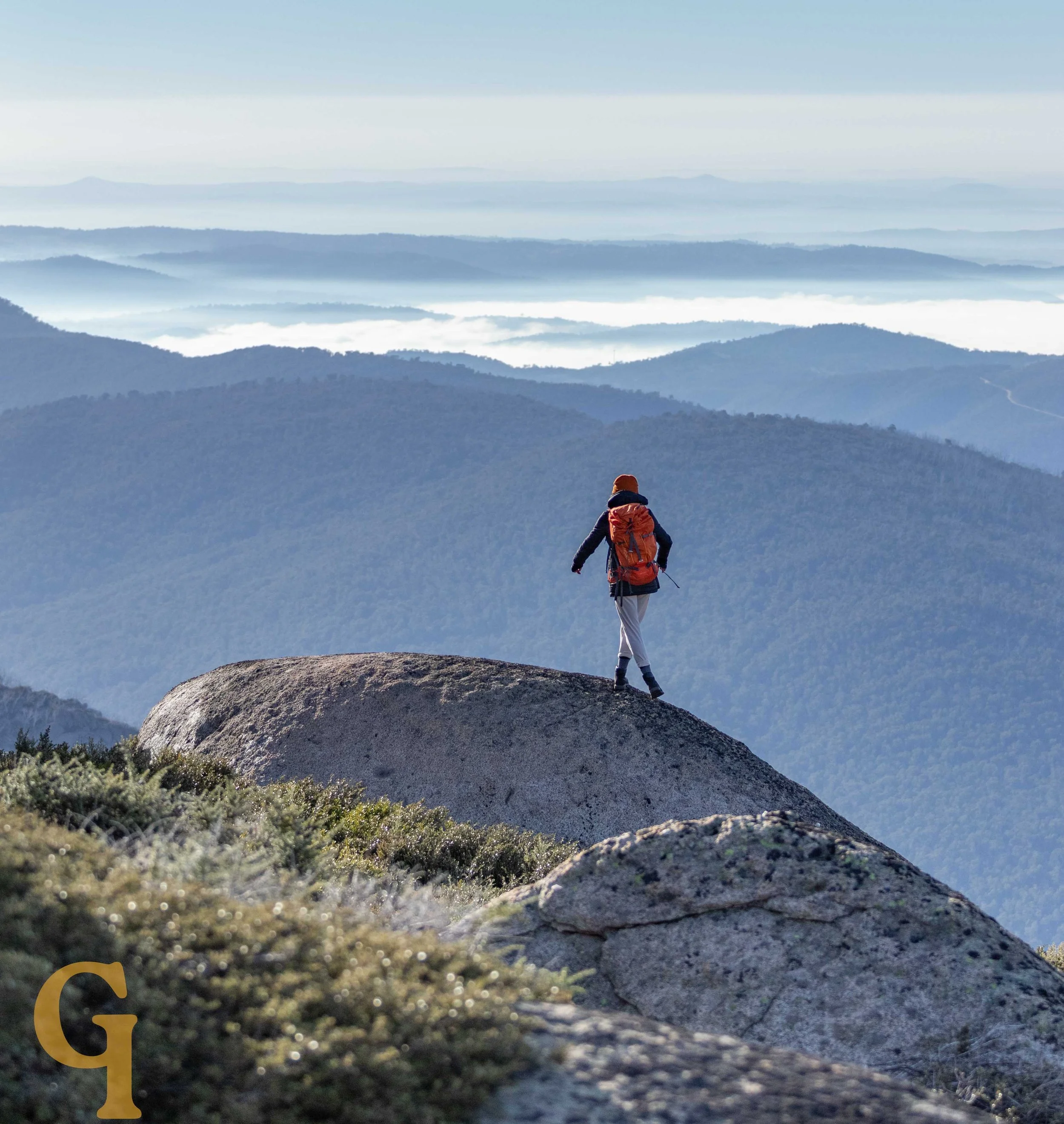 Person with a backpack hiking on a large rock on a mountain, with layers of mountain ridges and clouds in the background.