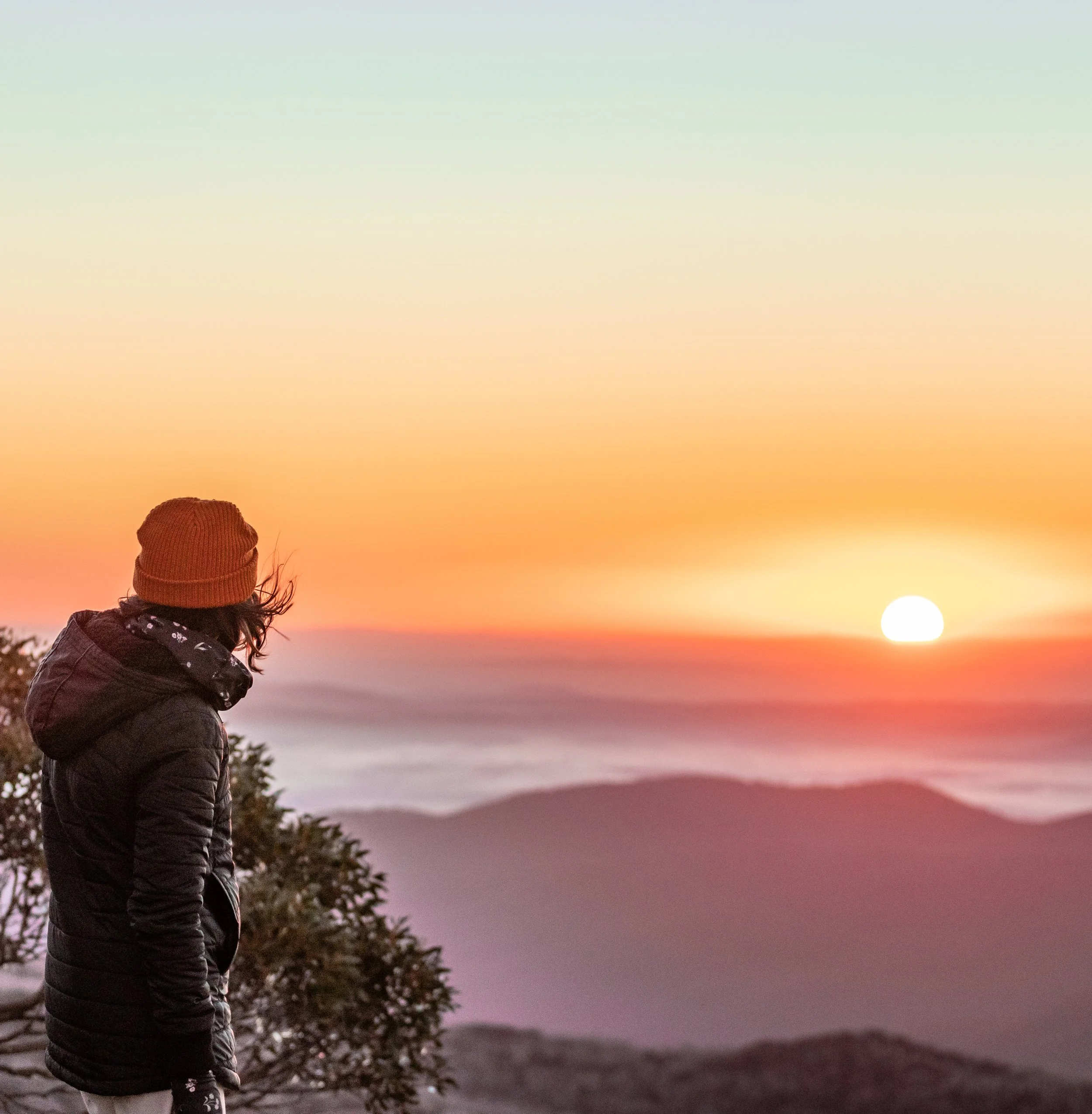 Person wearing a brown beanie and a black jacket looking at a sunset over mountains.