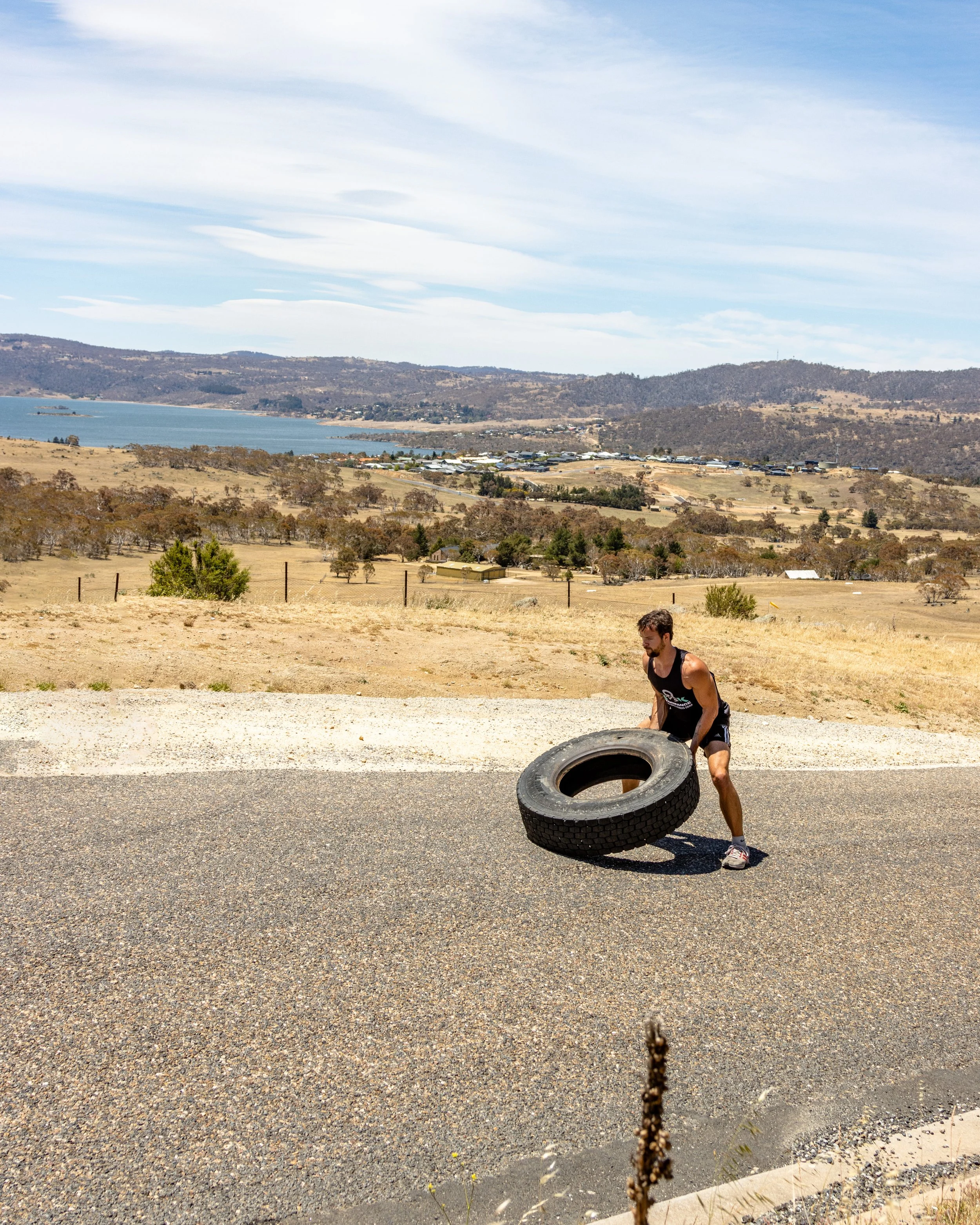 Man lifting a large tire outdoors on a gravel surface with rolling hills, a lake, and mountains in the background under a partly cloudy sky.