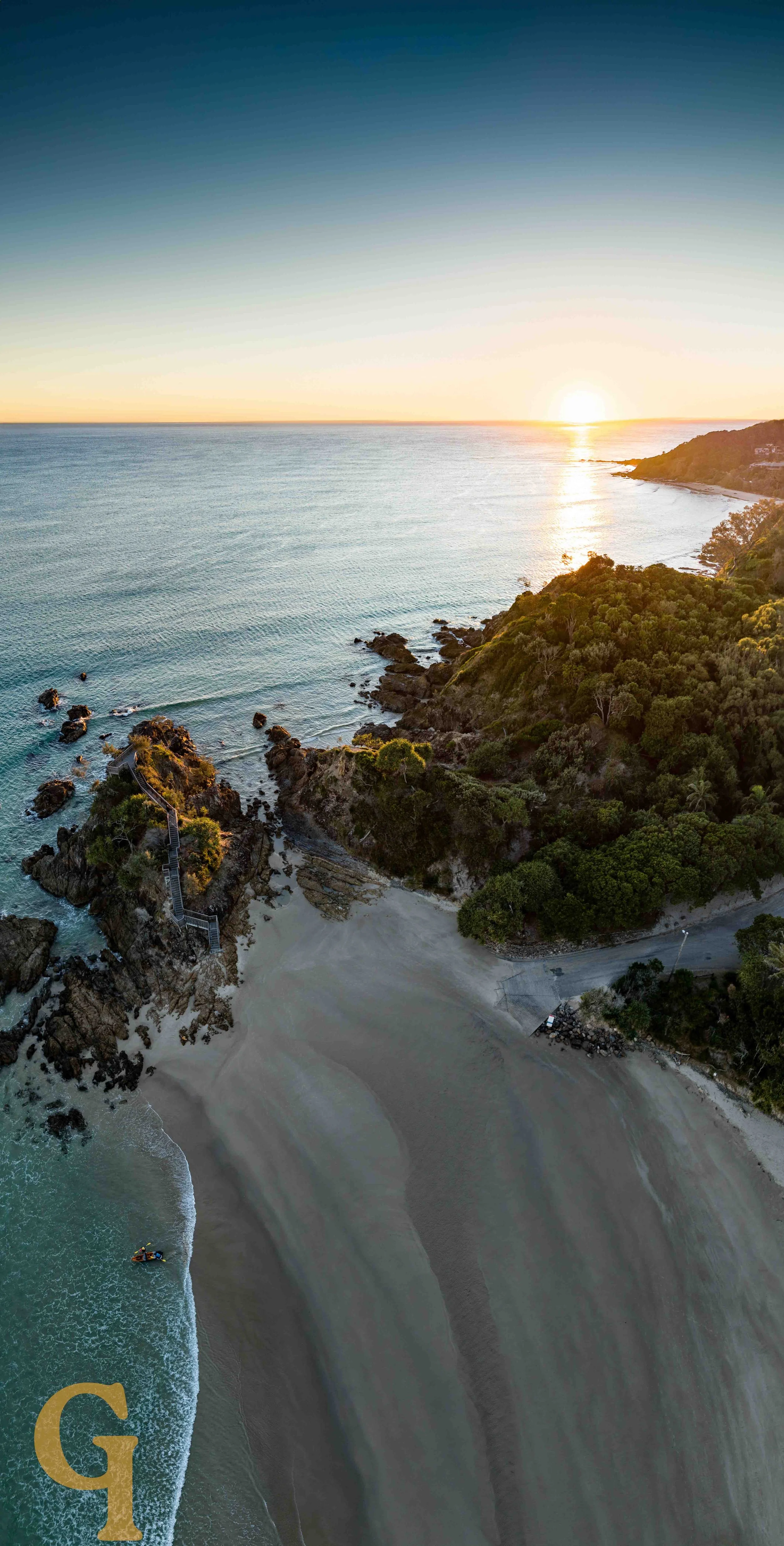 Aerial view of a sandy beach, rocky shoreline, and green hillside at sunrise, with a lighthouse on the hilltop.
