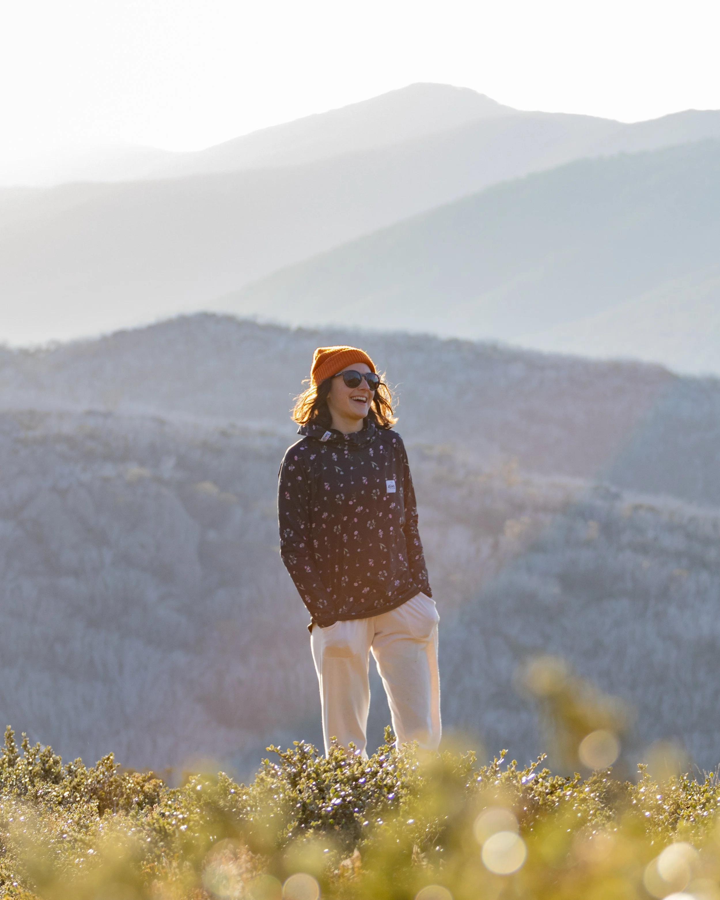 A woman standing outdoors in a mountainous landscape, wearing sunglasses, an orange beanie, a black jacket, and beige pants, smiling and enjoying the scenery.