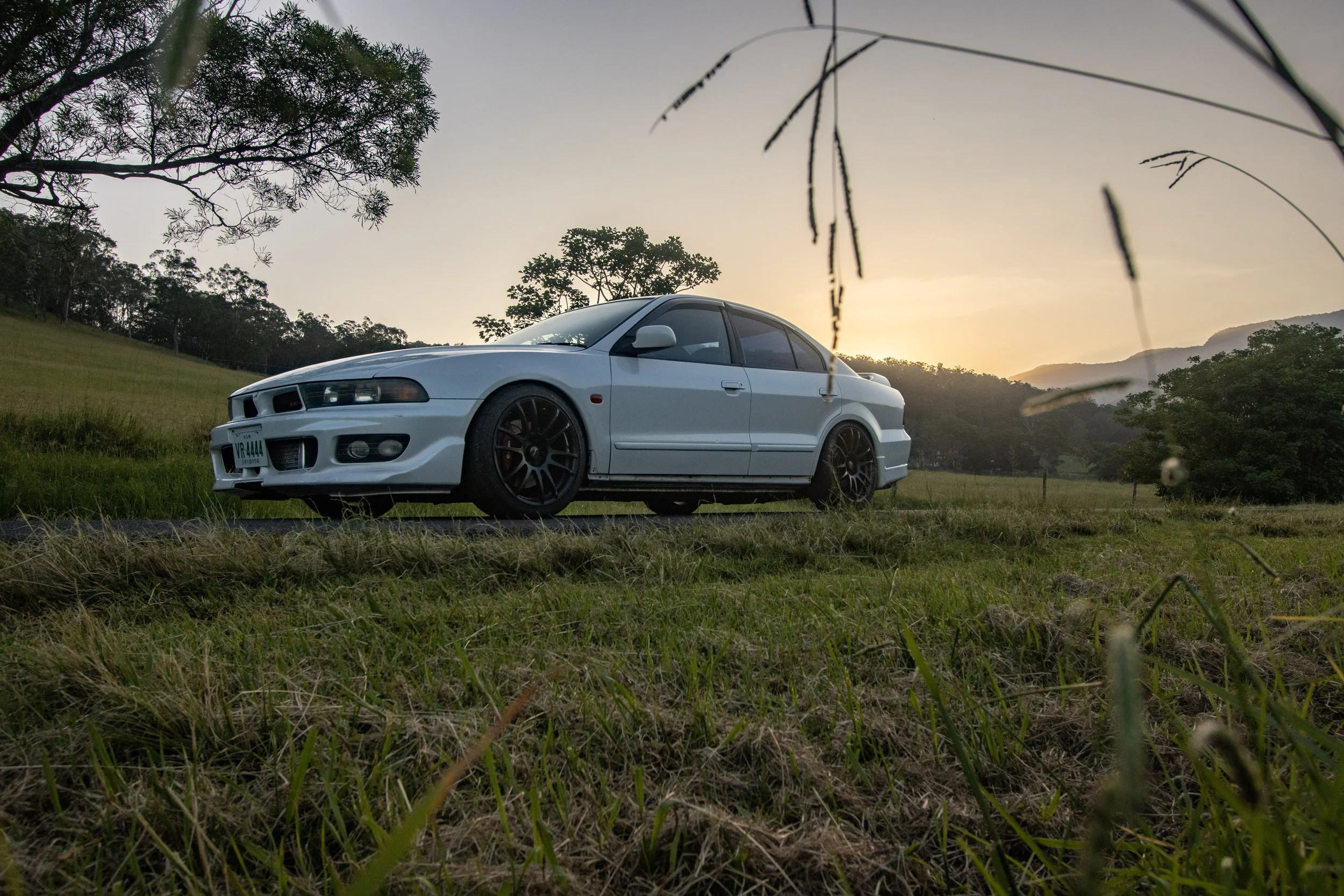A white sedan car parked on grass with trees and hills in the background during sunset.