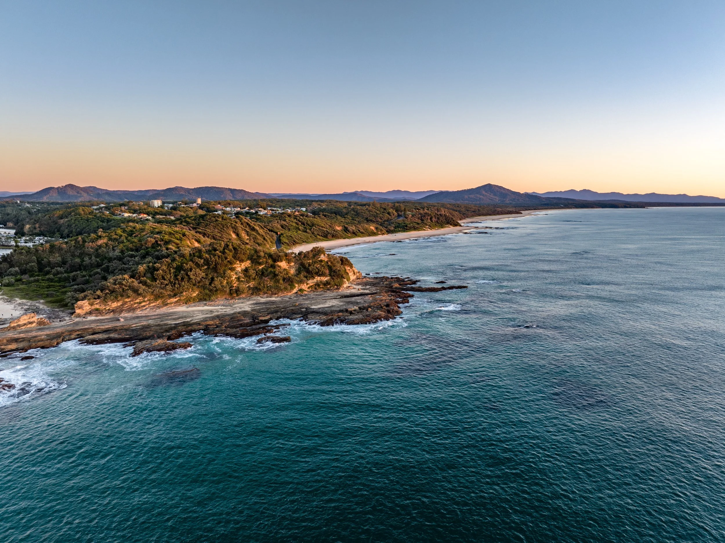 Aerial view of a coastline with rocky shores, sandy beaches, green forests, hills, and distant mountains at sunset.