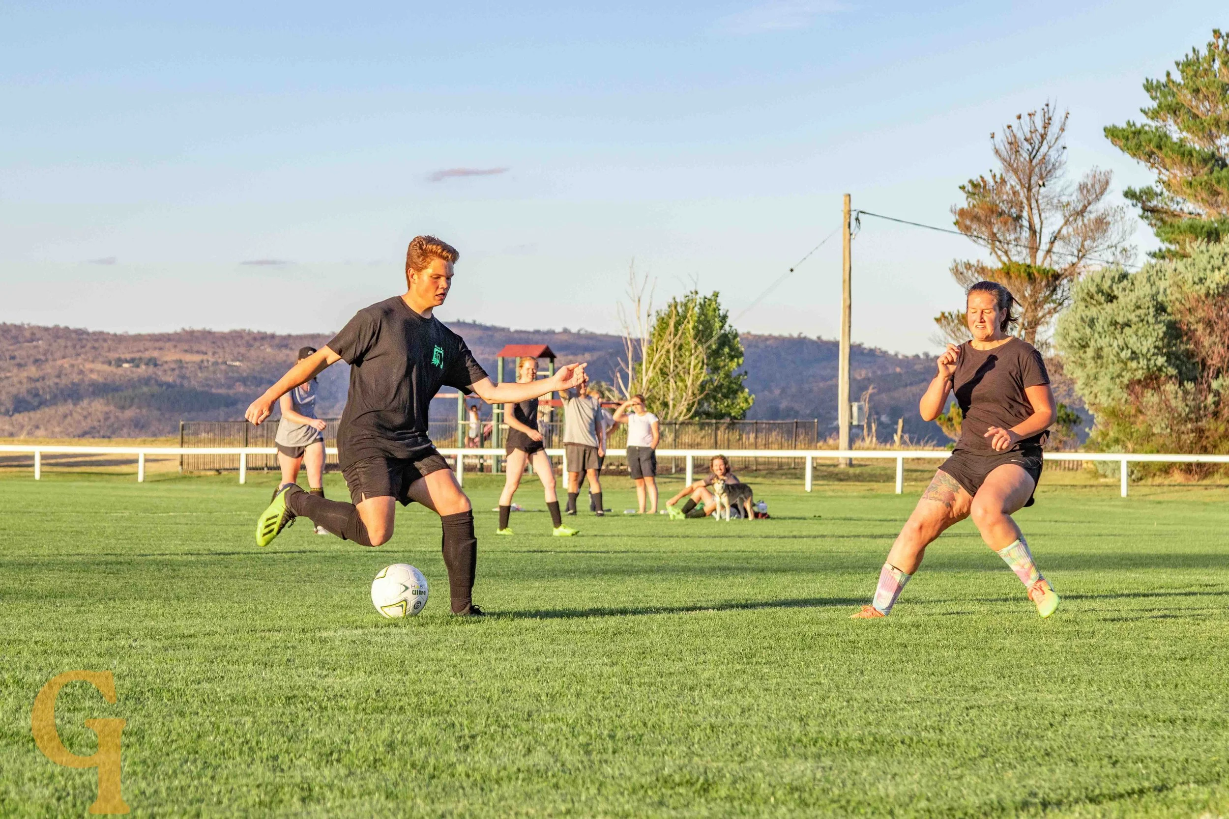 A group of people playing soccer on a grassy field, with two women actively competing for the ball, and others watching or relaxing in the background under clear skies.