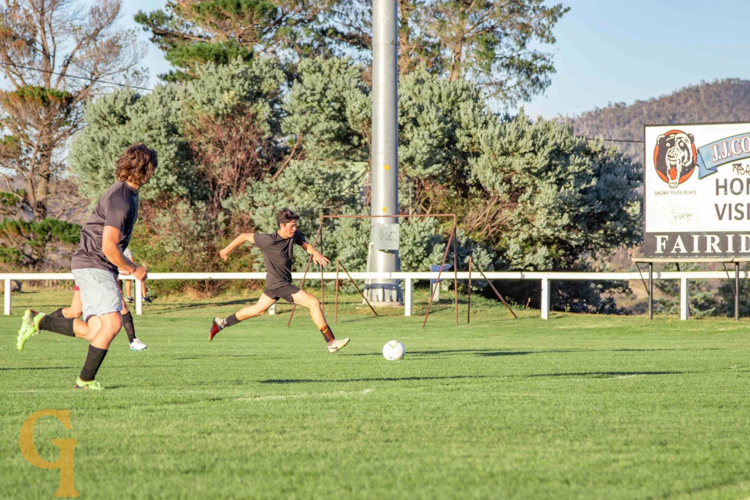 Two young men playing soccer on a grassy field with a goalpost in the background, surrounded by trees.