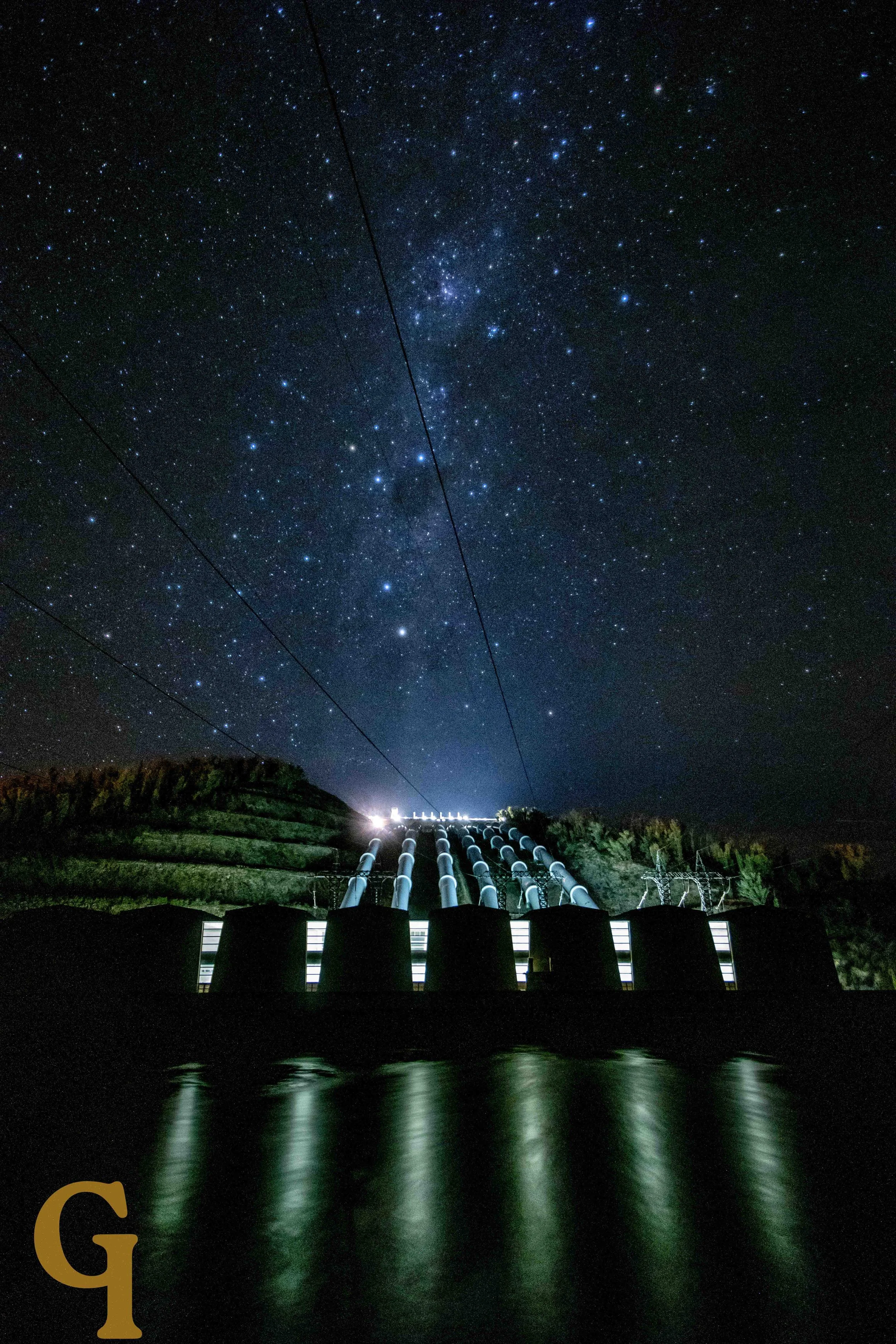 Nighttime view of a hydroelectric dam with water reflecting lights, under a starry sky with visible stars and the Milky Way galaxy.