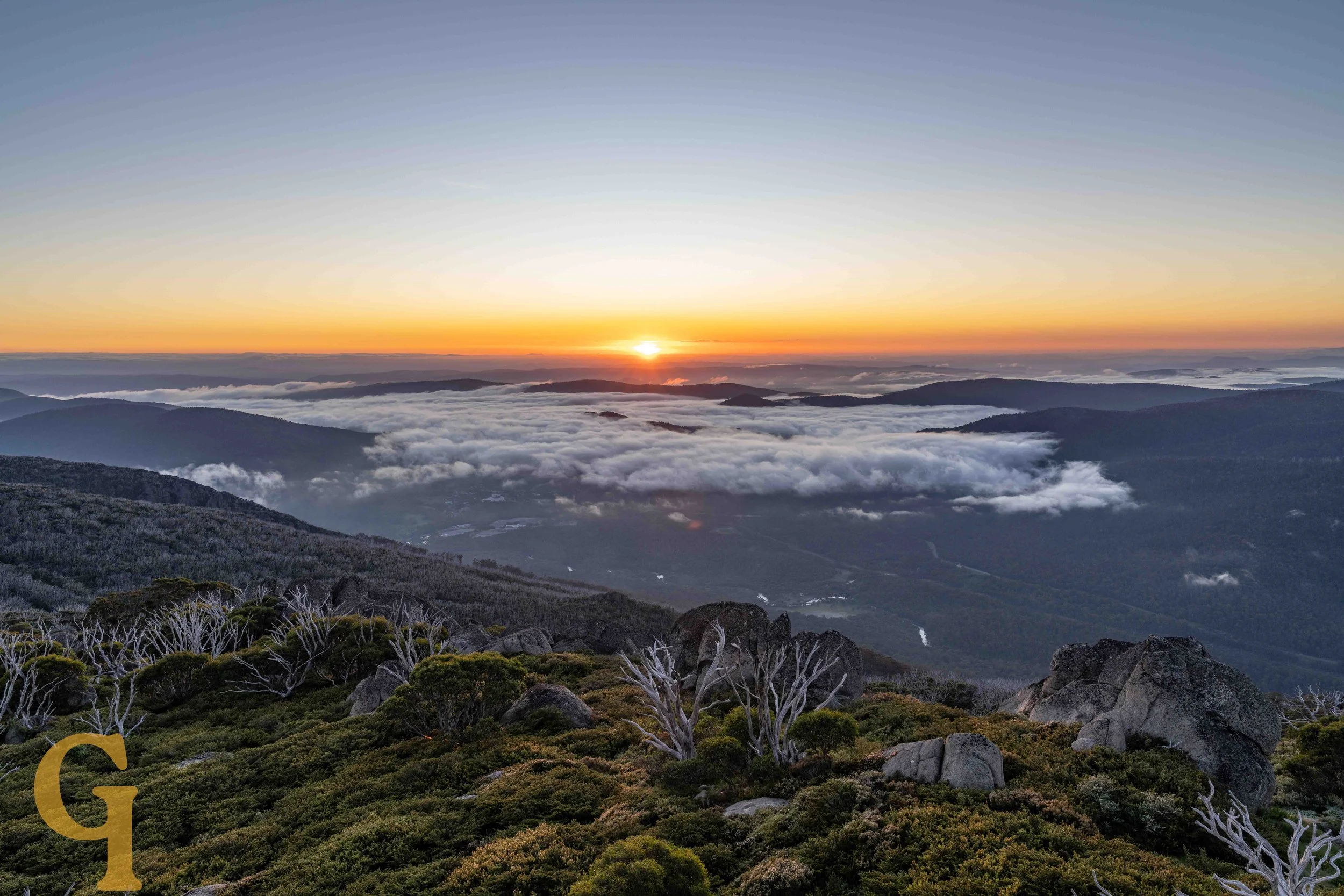 Sunrise over a mountain landscape with mist and clouds, rocky foreground with sparse trees and shrubs.