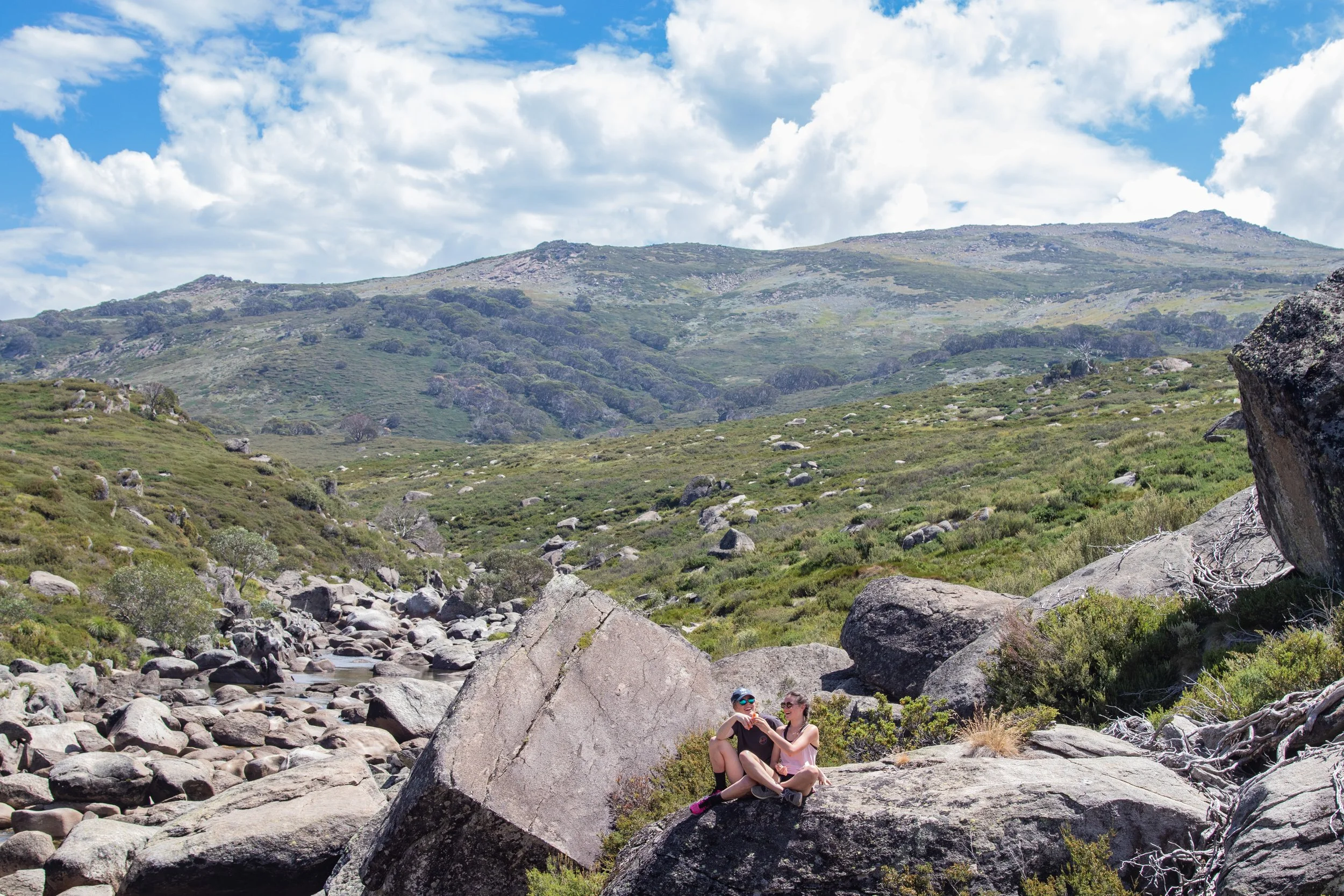 Two people sitting on a large rock in a mountainous landscape with green hills, rocks, and a partly cloudy sky.