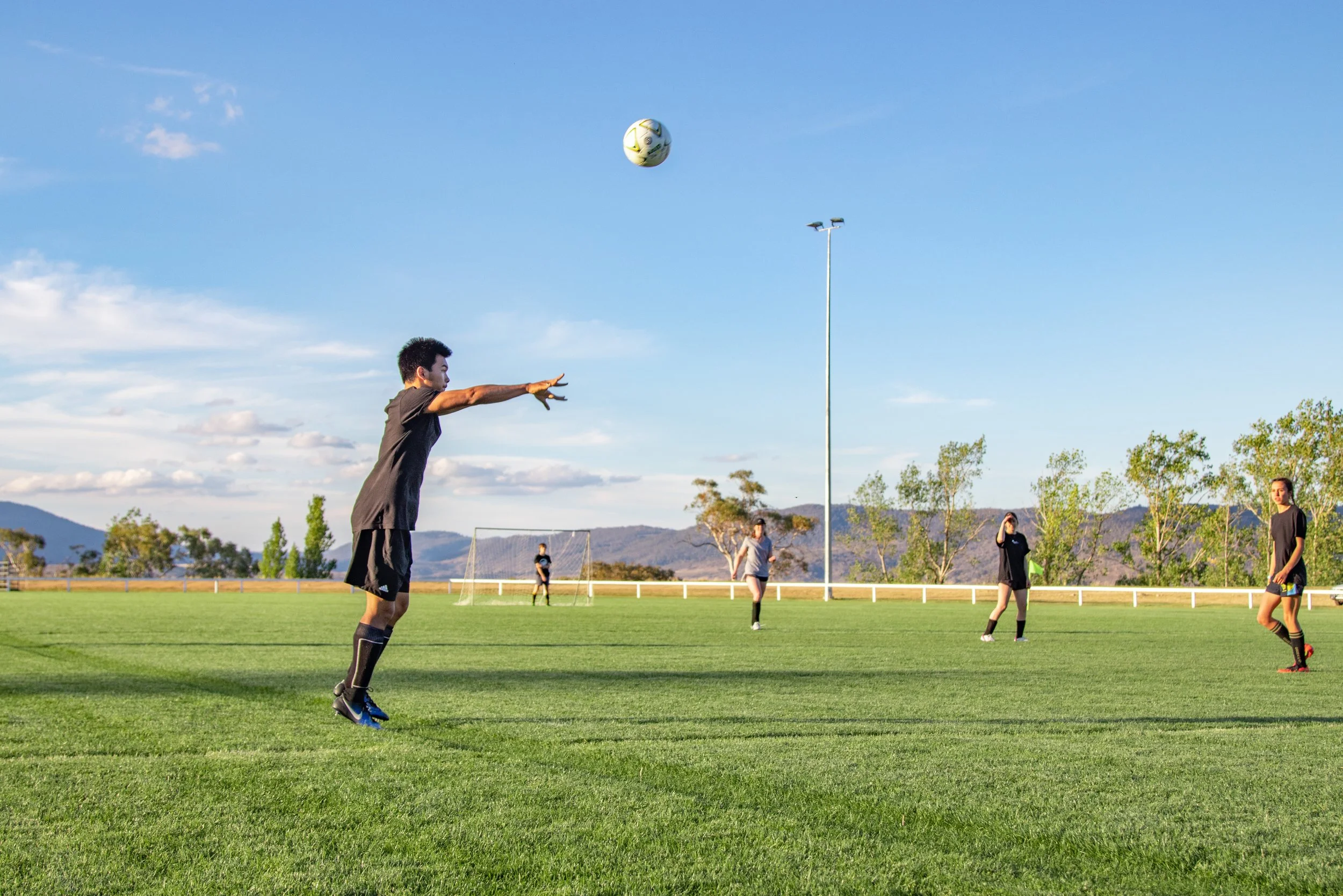 A group of women and a man playing soccer on a grassy field under a blue sky with some clouds, with mountains and trees in the background. One man is about to catch a soccer ball.