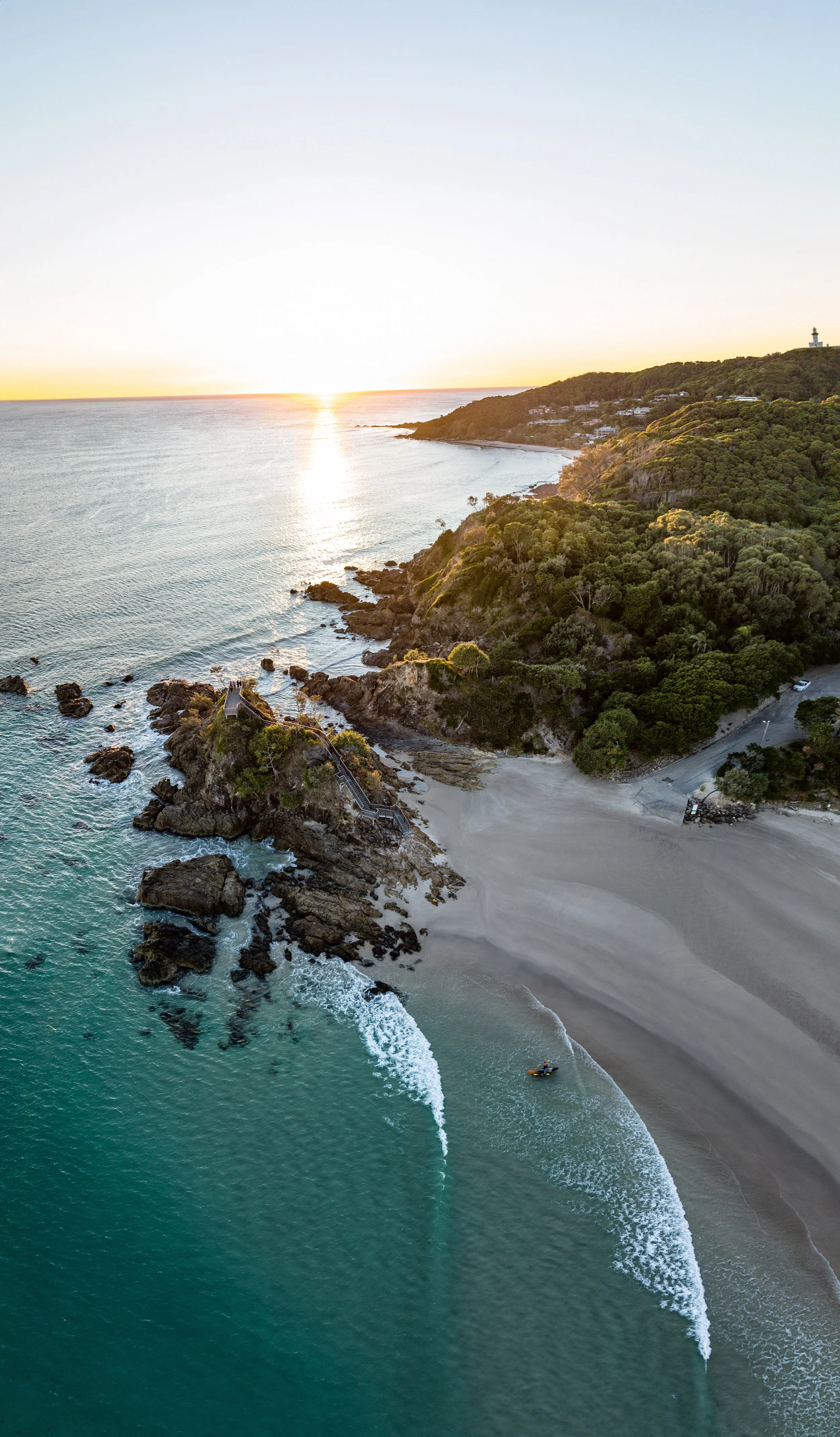 Aerial view of a sandy beach, rocky shoreline, and green hillside at sunset, with a lighthouse on the hilltop.