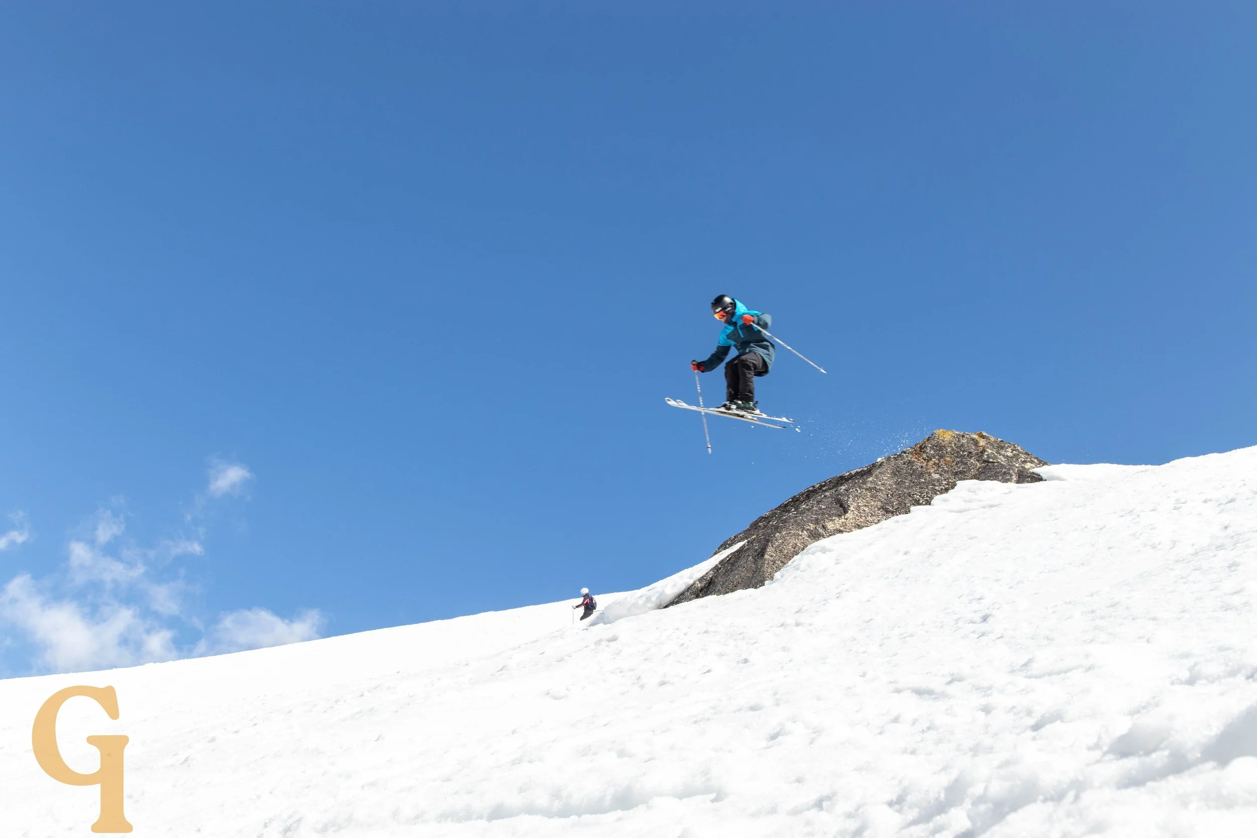 A skier in a blue jacket and black pants jumping off a snow-covered slope with a clear blue sky in the background.