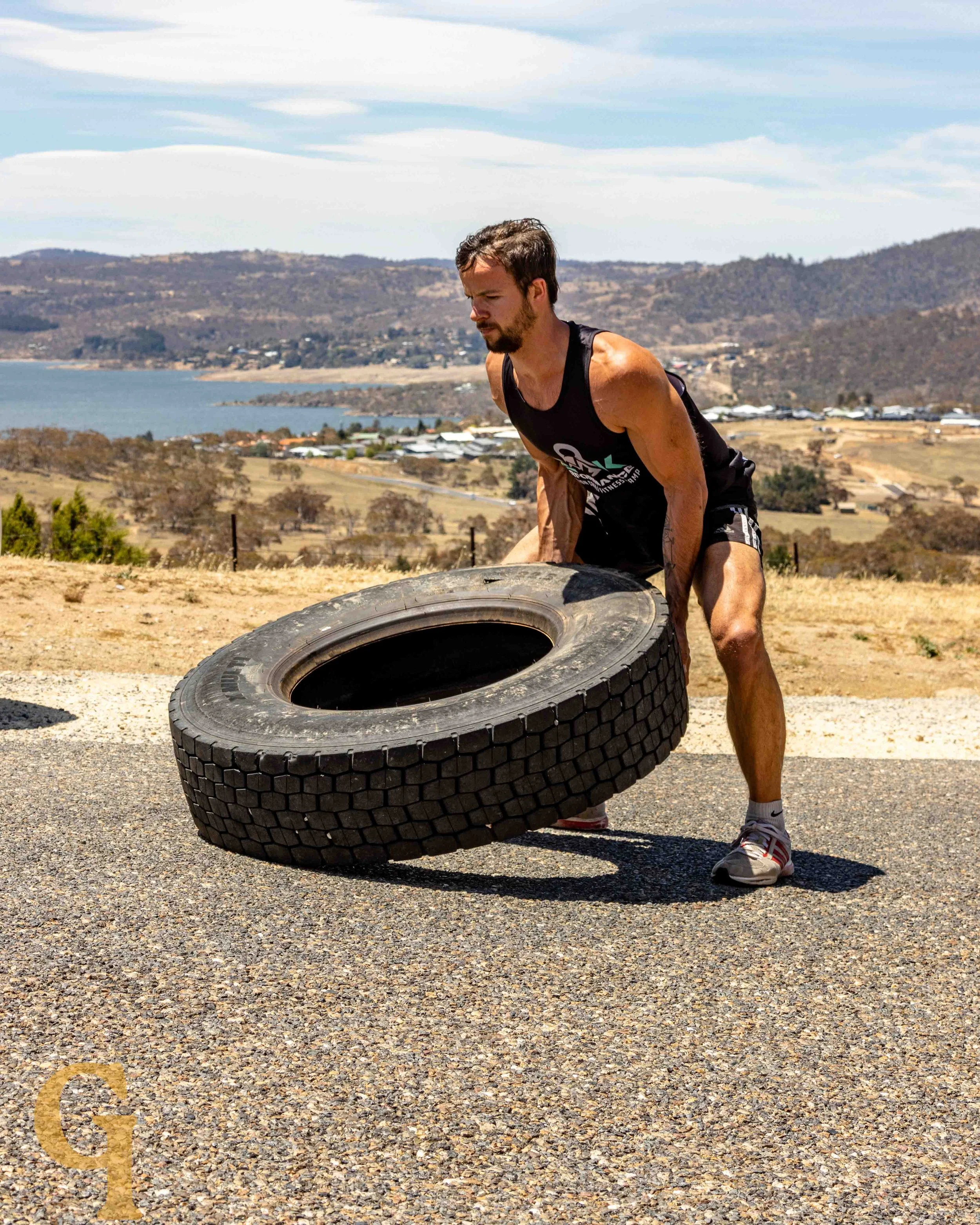 A man in athletic attire lifting a large tire outdoors on a sunny day with hills, a lake, and a rural landscape in the background.