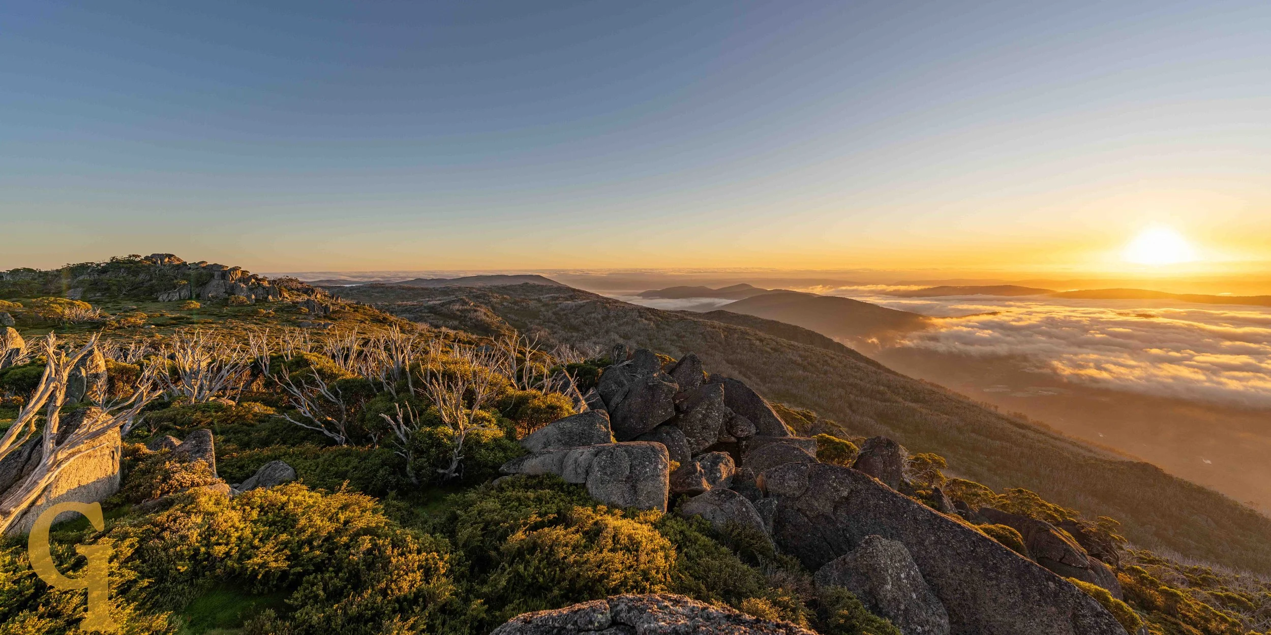 Sunrise over a mountain landscape with rocky terrain, sparse trees, and a sea of clouds in the distance.