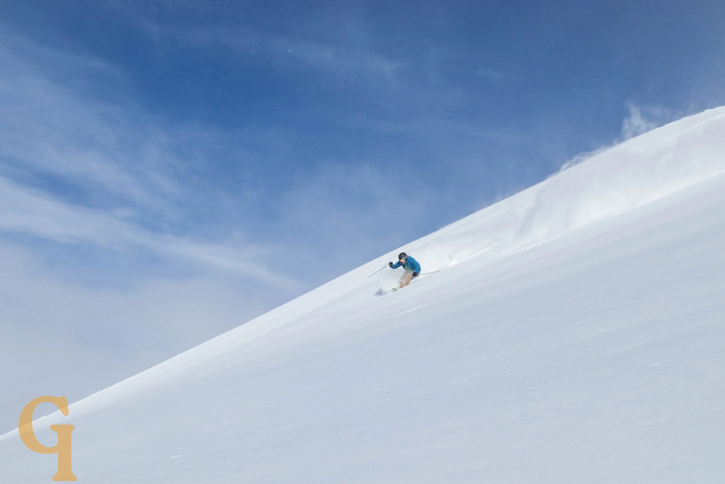 A skier in blue and black gear skiing down a snow-covered slope on a clear, sunny day.