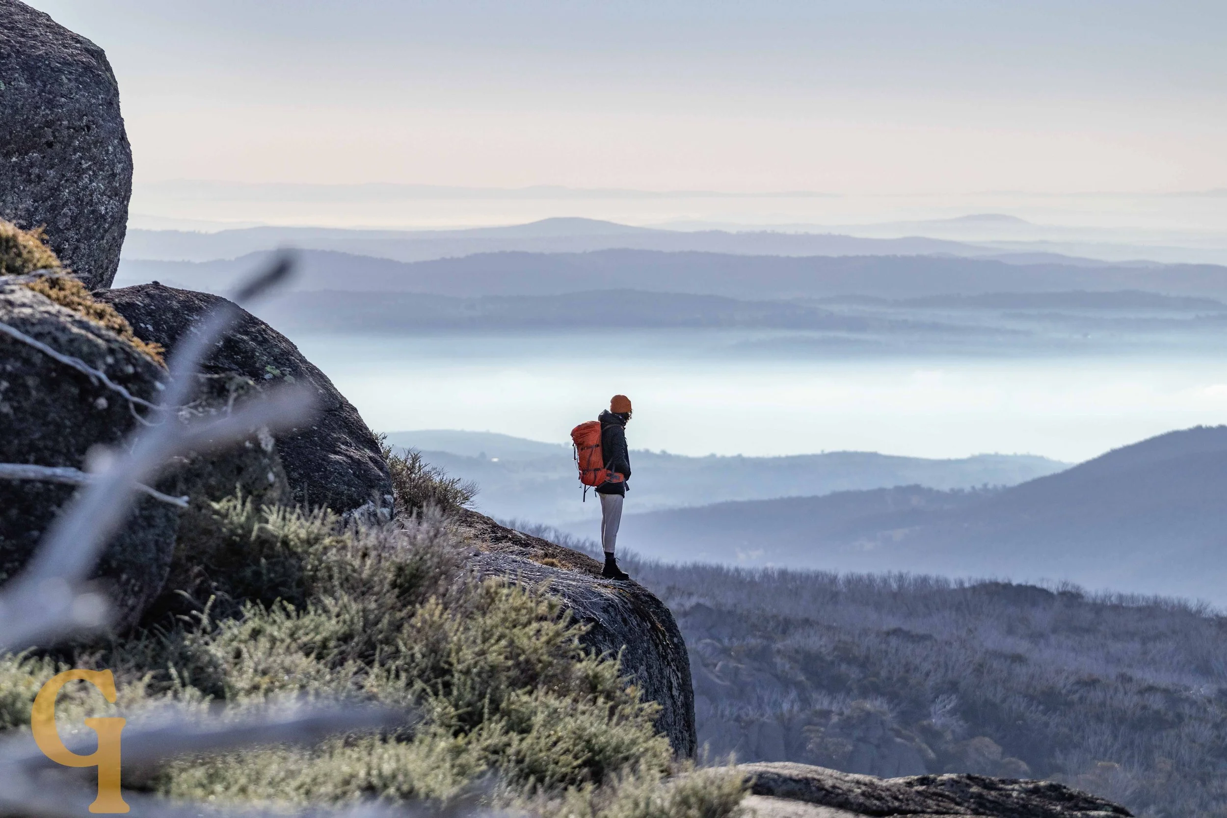 A hiker with a backpack standing on a rocky mountain ledge overlooking a misty, layered landscape of hills and water bodies.